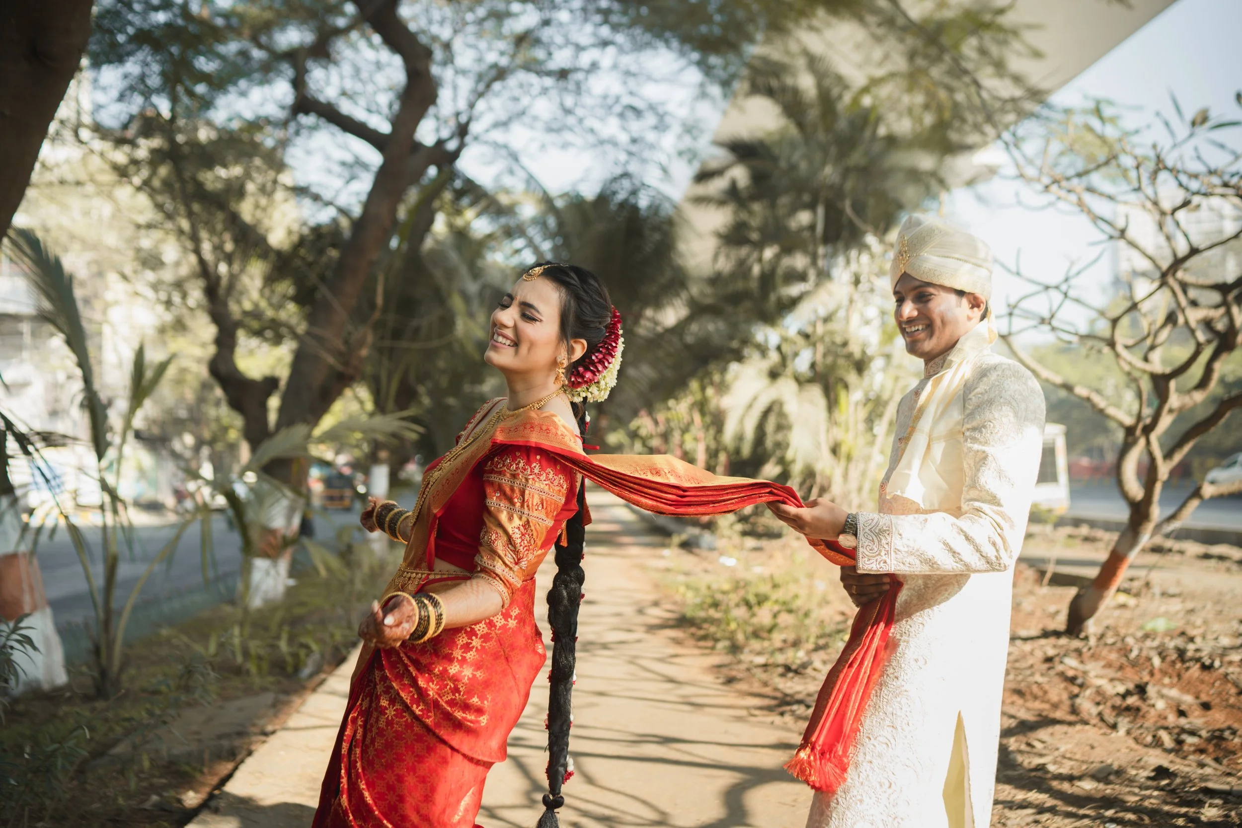 Indian couple in traditional wedding attire, with the groom pulling the bride's dupatta, smiling and walking outdoors on a sunny day.