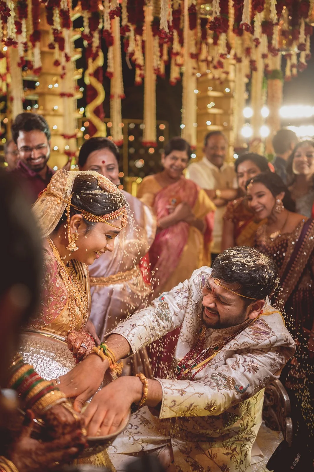 An Indian wedding ceremony showing the bride and groom participating in a ritual surrounded by family and friends at night, decorated with flowers and string lights.