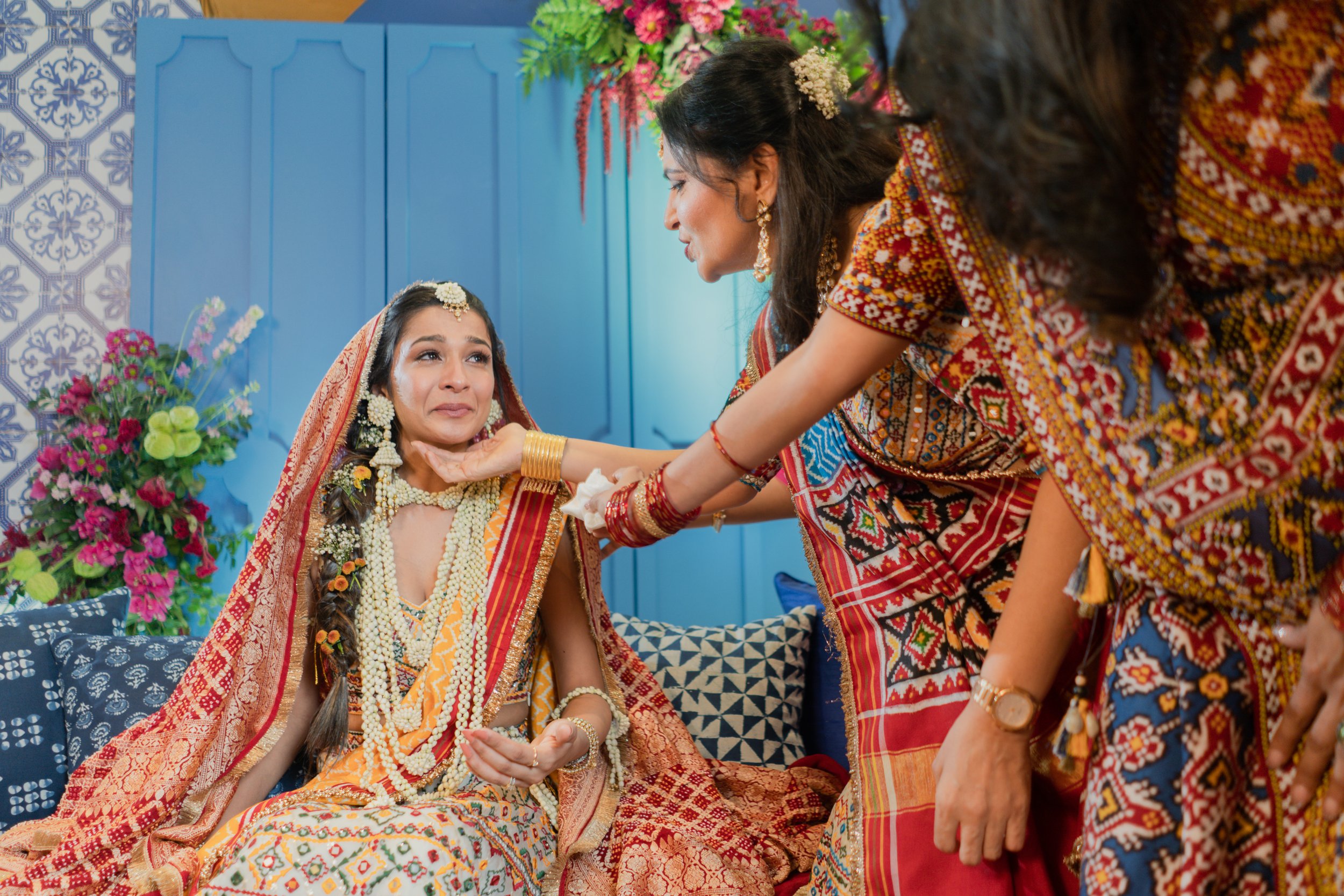 A woman dressed in traditional South Asian wedding attire is seated, crying, as another woman in colorful attire washes her feet. The scene is decorated with vibrant flowers and patterned fabrics.