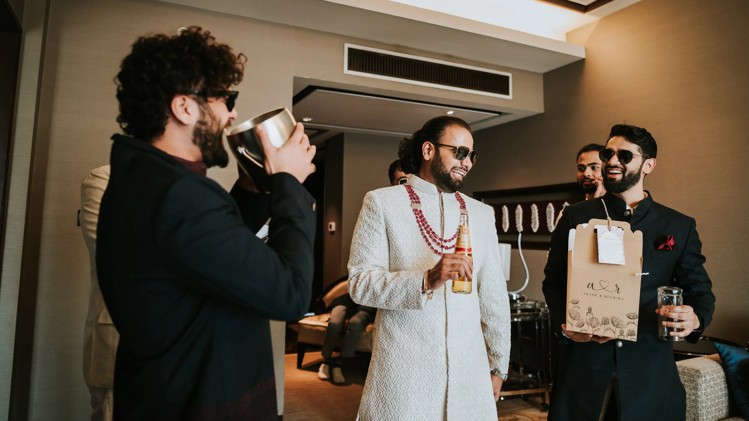Group of men at a celebration, smiling and holding drinks, in a modern indoor setting, with one man holding a gift bag and another drinking from a cup, all wearing formal or traditional attire and sunglasses.