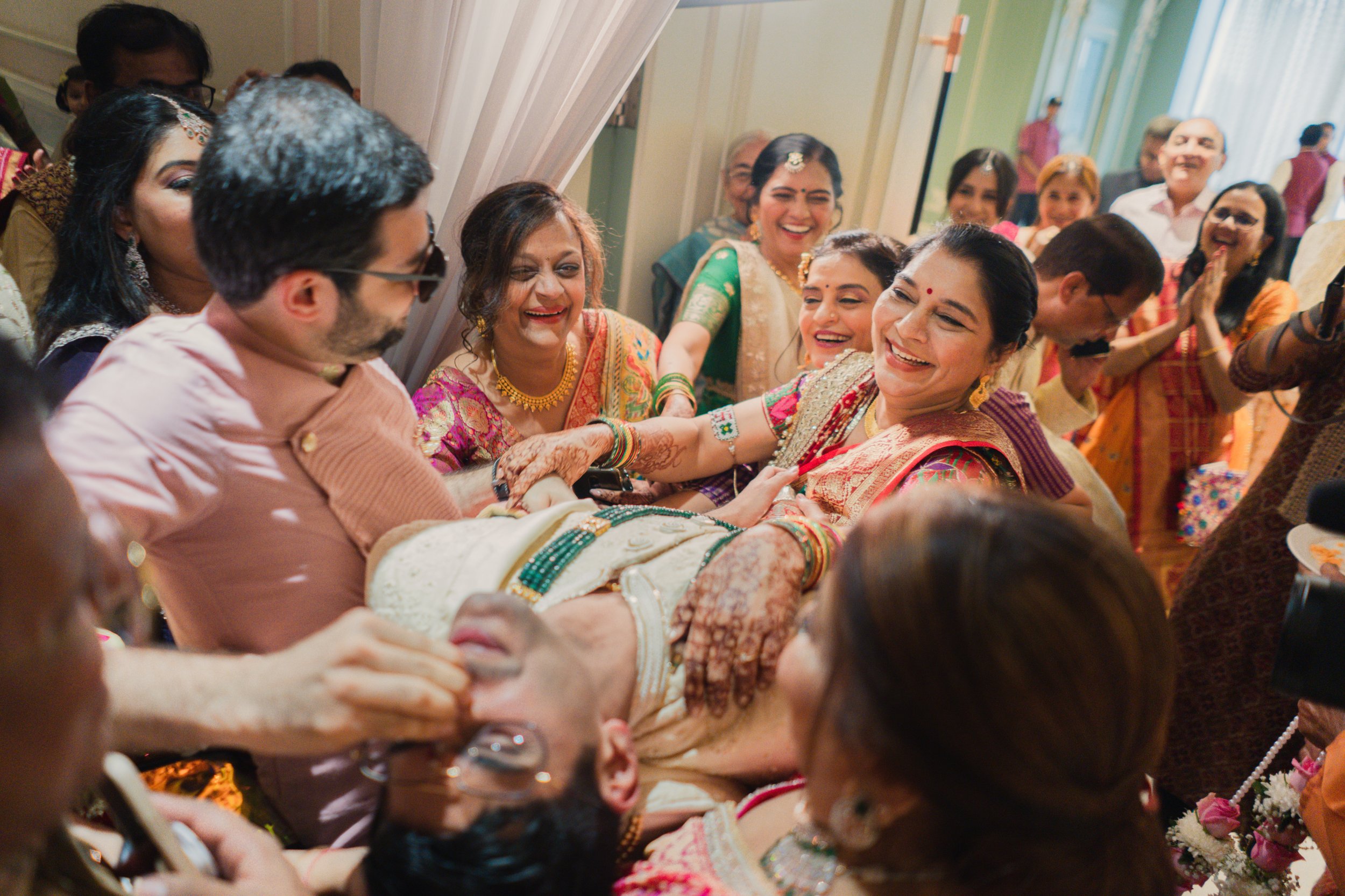 Group of people celebrating around a woman in traditional Indian attire, who is being lifted or supported by others, smiling and laughing during a festive occasion.