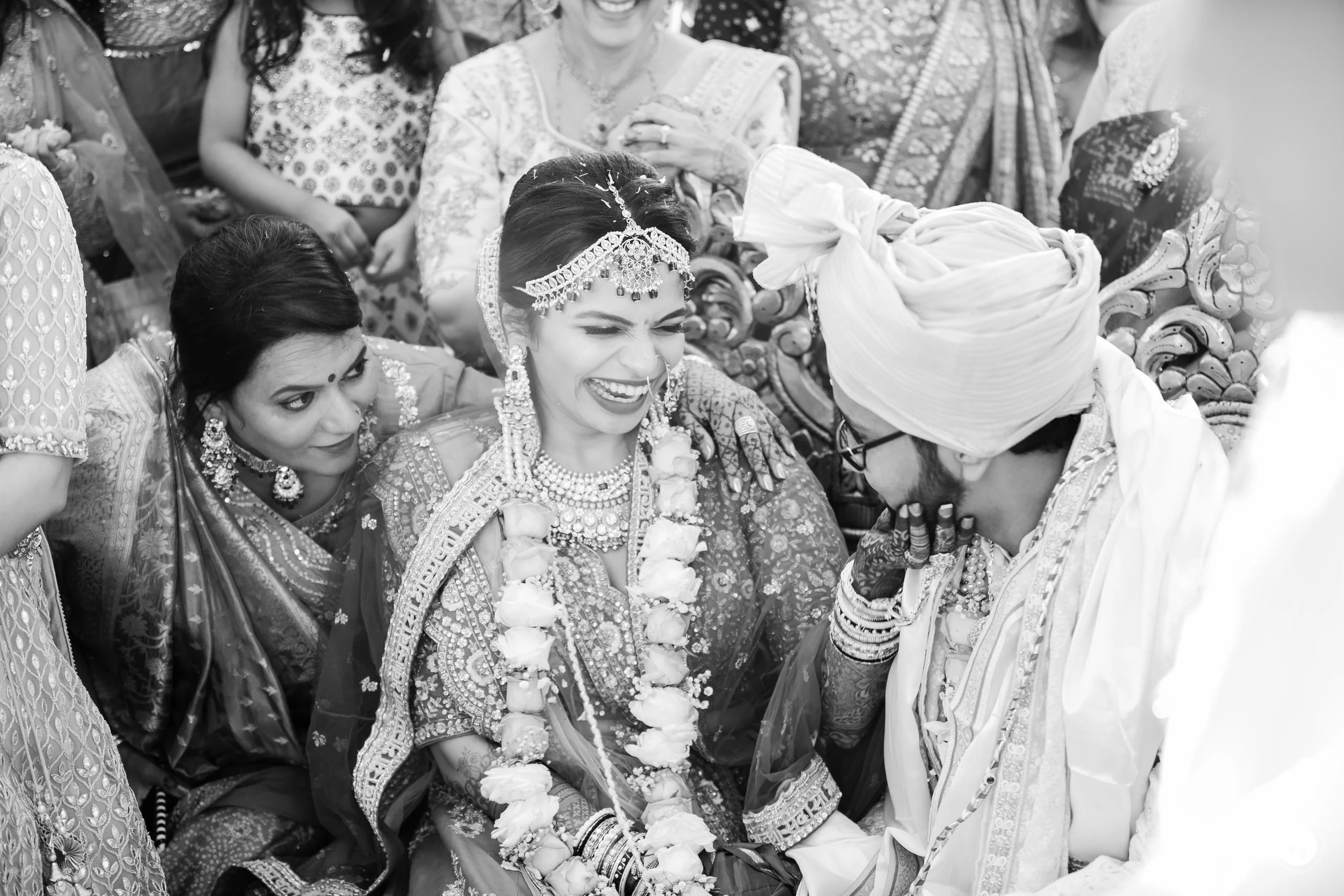 A bride smiling happily during her traditional Indian wedding ceremony, surrounded by women and a groom wearing a turban, in a black and white photo.