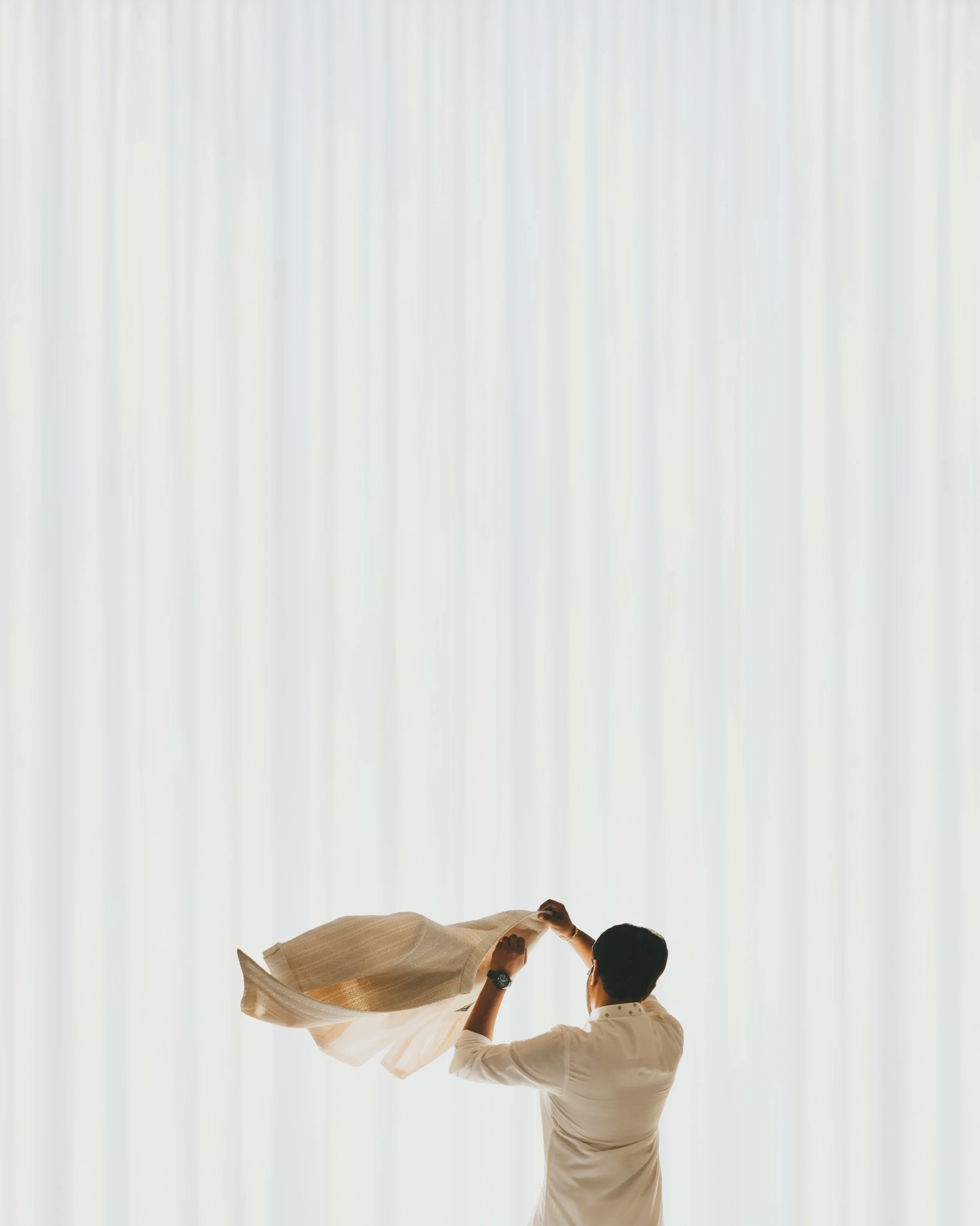 Man standing with back to camera, holding a beige curtain or fabric, in front of a large window with white curtains.