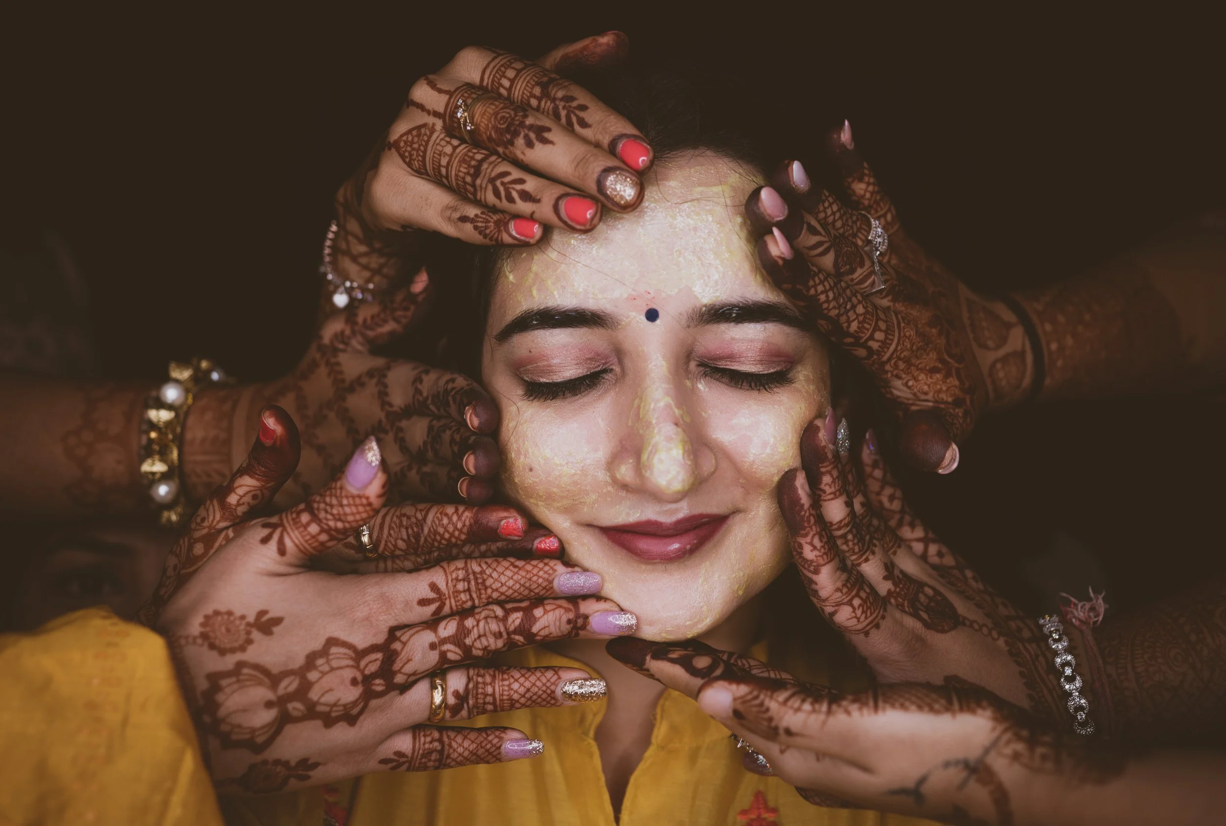A woman with closed eyes receives a traditional henna tattoo session, with multiple hands decorating her face. The hands are adorned with jewelry and have intricate henna designs.