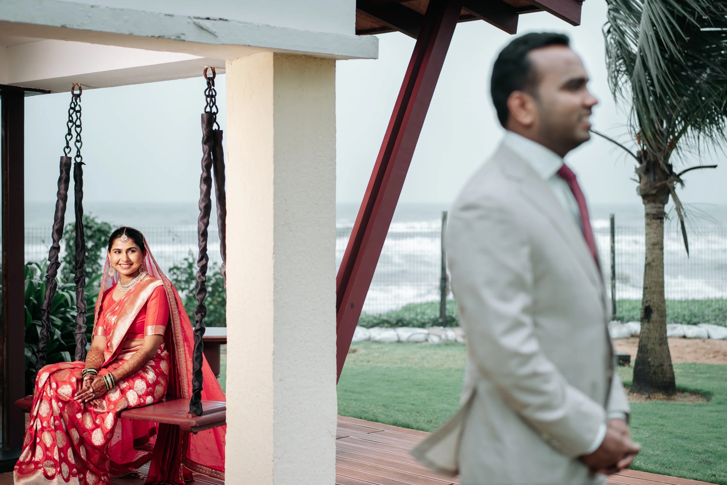 A woman in traditional Indian attire sitting on a swing, smiling, with a man in a white suit standing in the background outdoors near the beach.