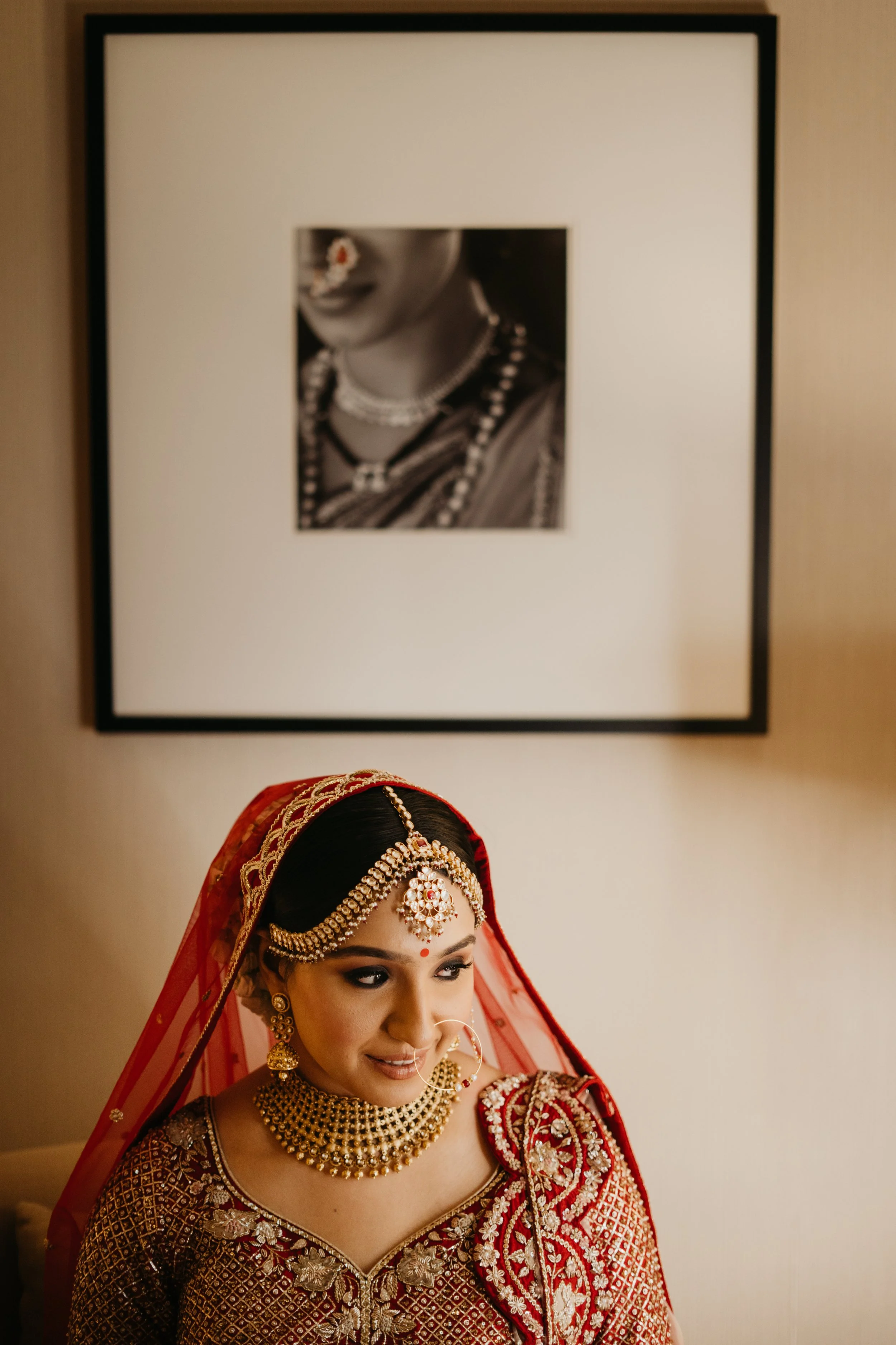 A woman dressed in traditional Indian bridal attire with jewelry, earrings, forehead ornament, and red veil, smiling softly, sitting indoors with framed black-and-white photograph of a woman in jewelry on the wall behind her.