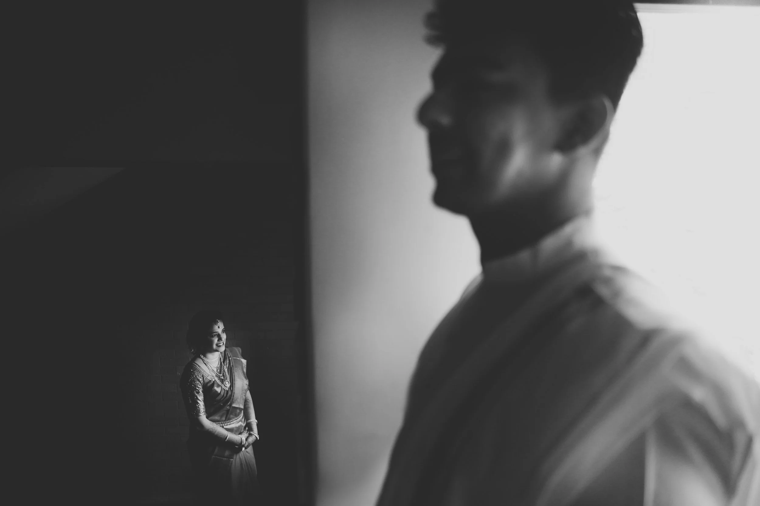Black and white photo of a woman in traditional Indian attire smiling at a man whose face is partially visible, both standing indoors.
