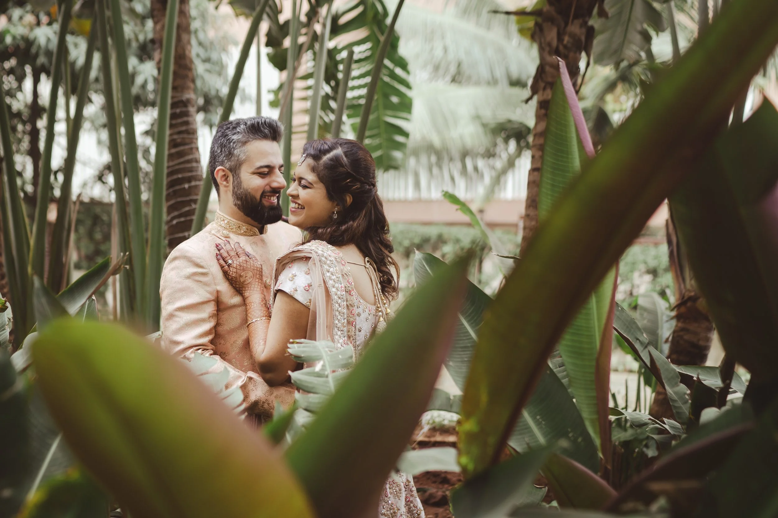 A joyful couple dressed in traditional Indian wedding attire smiling and touching foreheads in a lush, green tropical setting surrounded by large leaves and palm trees.