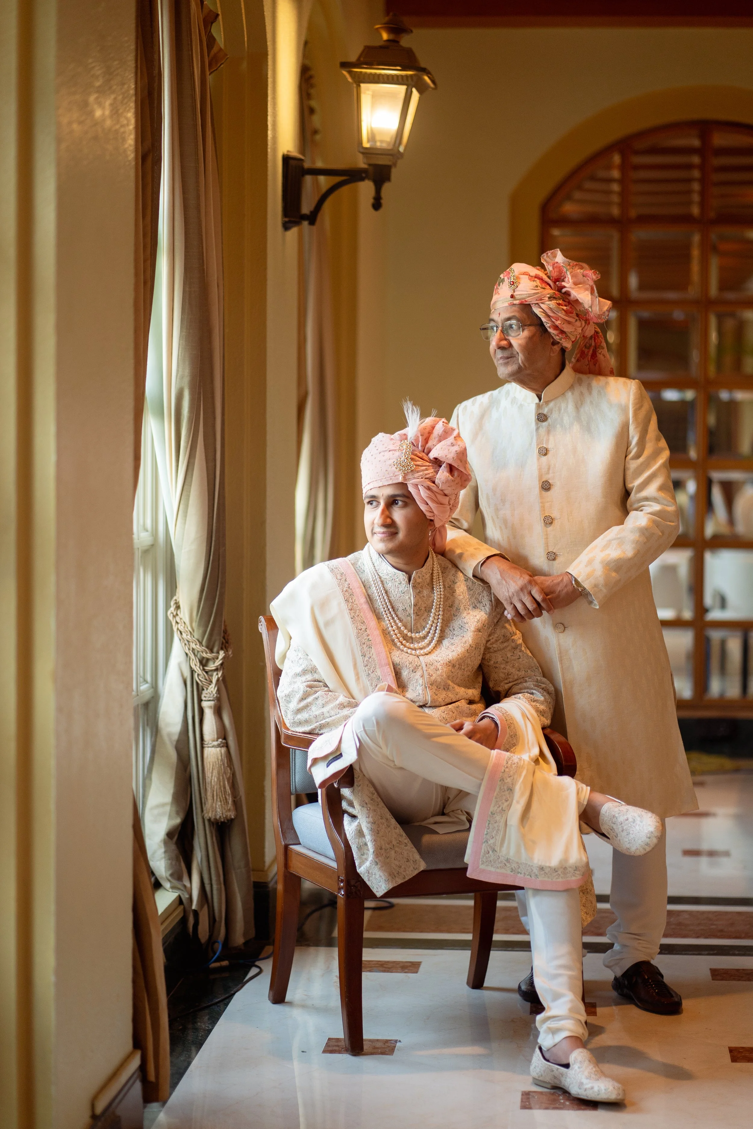 Two men dressed in traditional Indian wedding attire, one seated and one standing behind, in a well-lit room with beige walls, curtains, and a wooden partition.