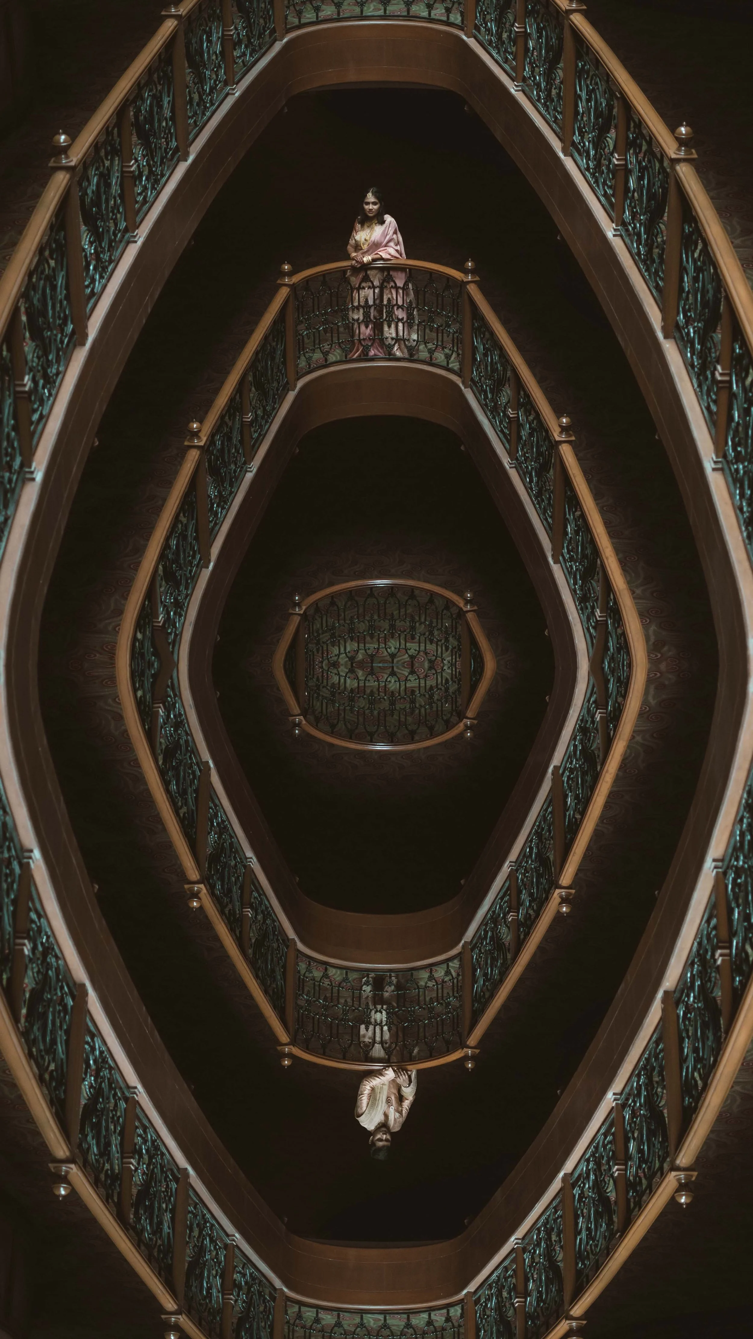 A woman in traditional attire standing on a balcony of a spiral staircase with ornate railing, viewed from above.