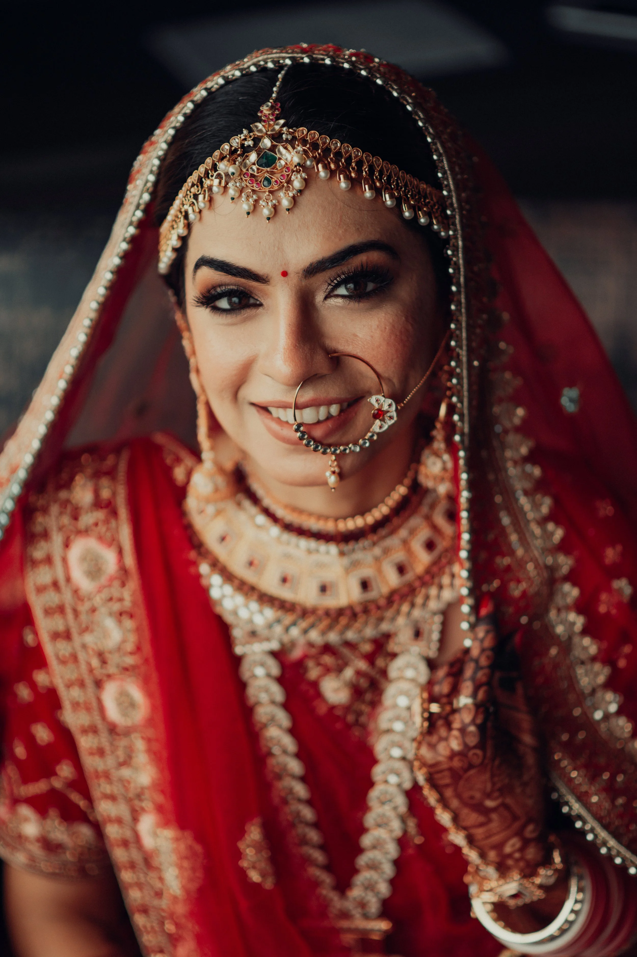 A woman dressed in traditional Indian bridal attire, wearing red and gold jewelry, including necklaces, earrings, a nose ring, and a headpiece, with a red veil and intricate embroidery on her clothing.