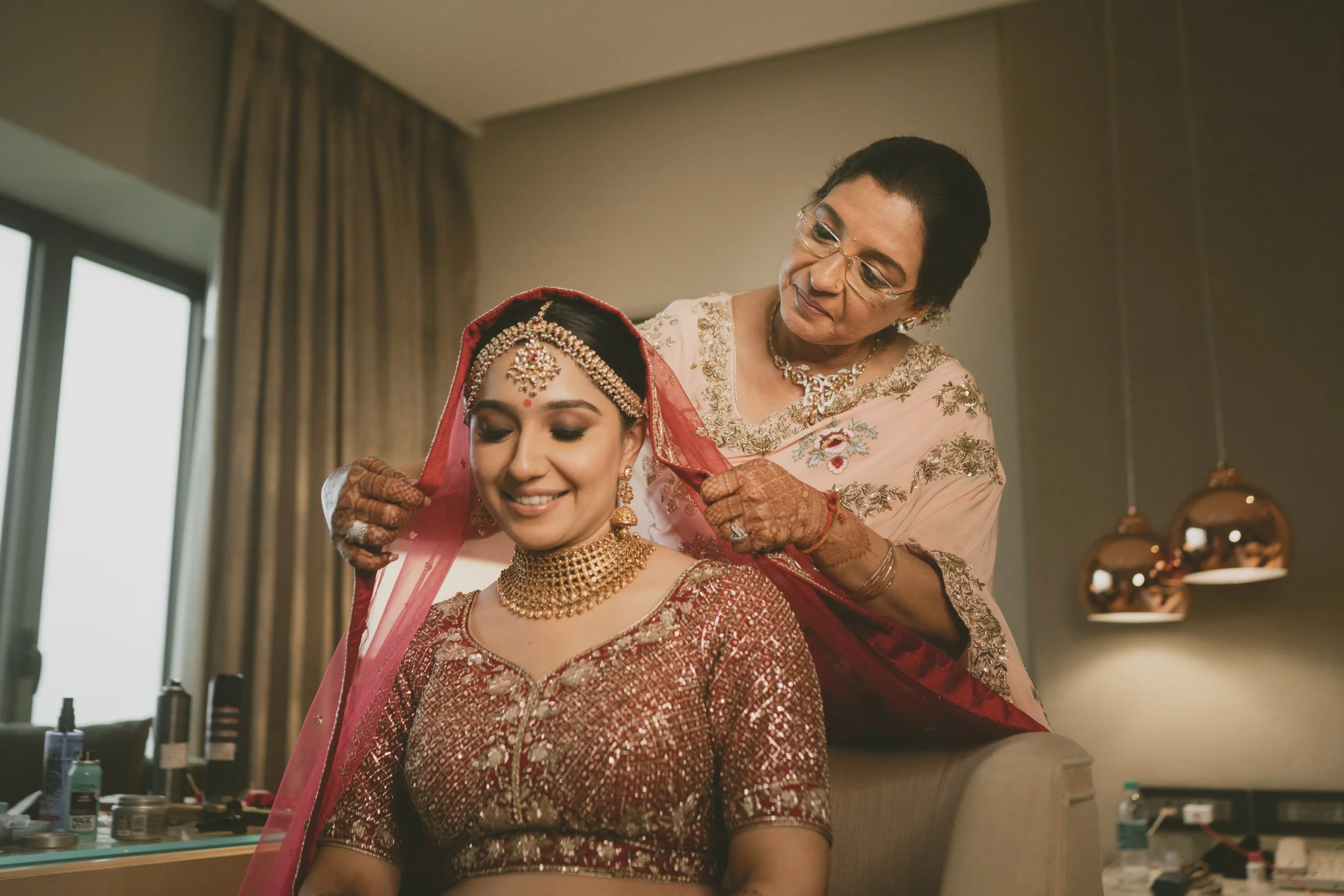 A bride dressed in traditional Indian wedding attire smiling as an older woman adjusts her pink veil inside a hotel room.
