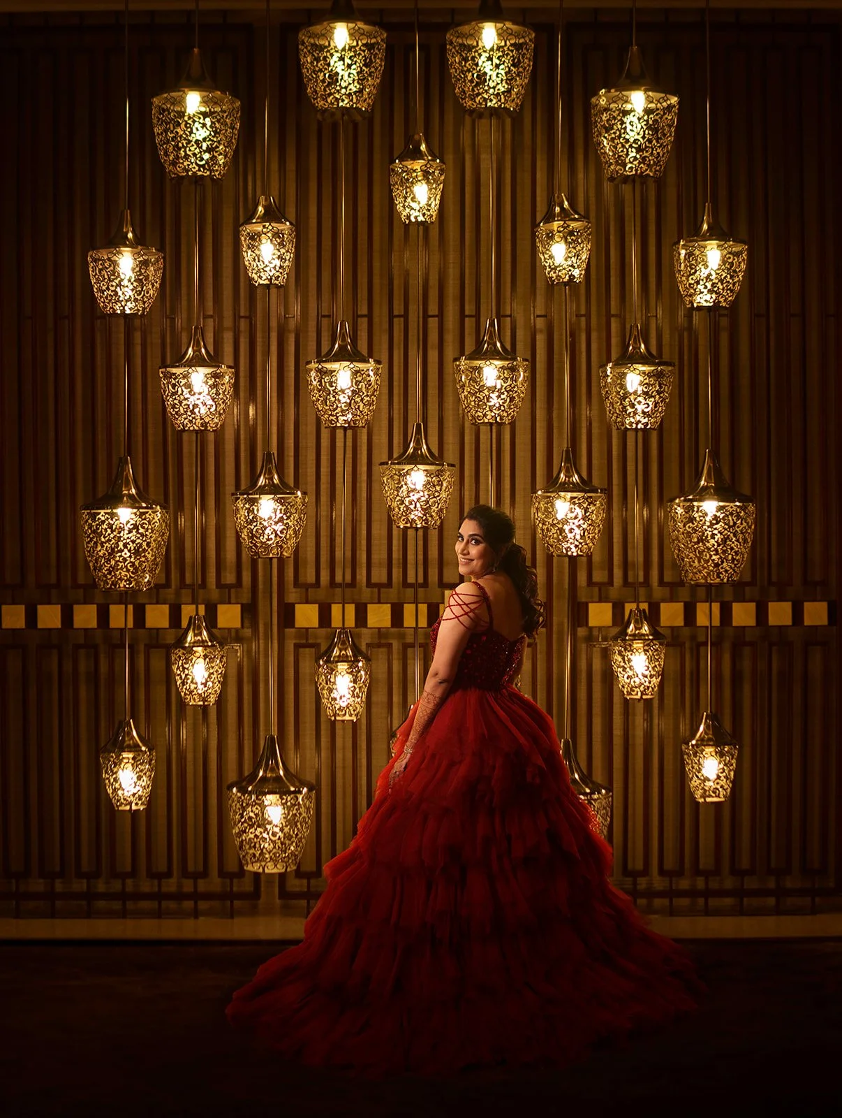 A woman in a red gown standing in front of an ornate, illuminated wall with pendant lights.