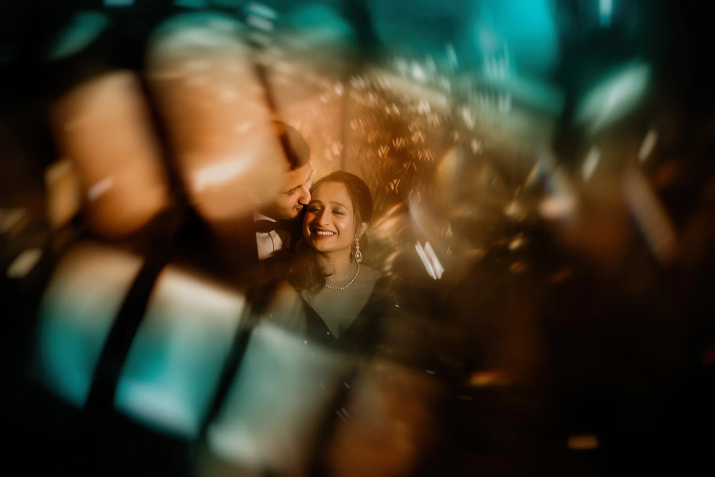 A couple dressed in formal attire, smiling and sharing an intimate moment at a party or celebration, with blurred warm lighting and reflections around them.