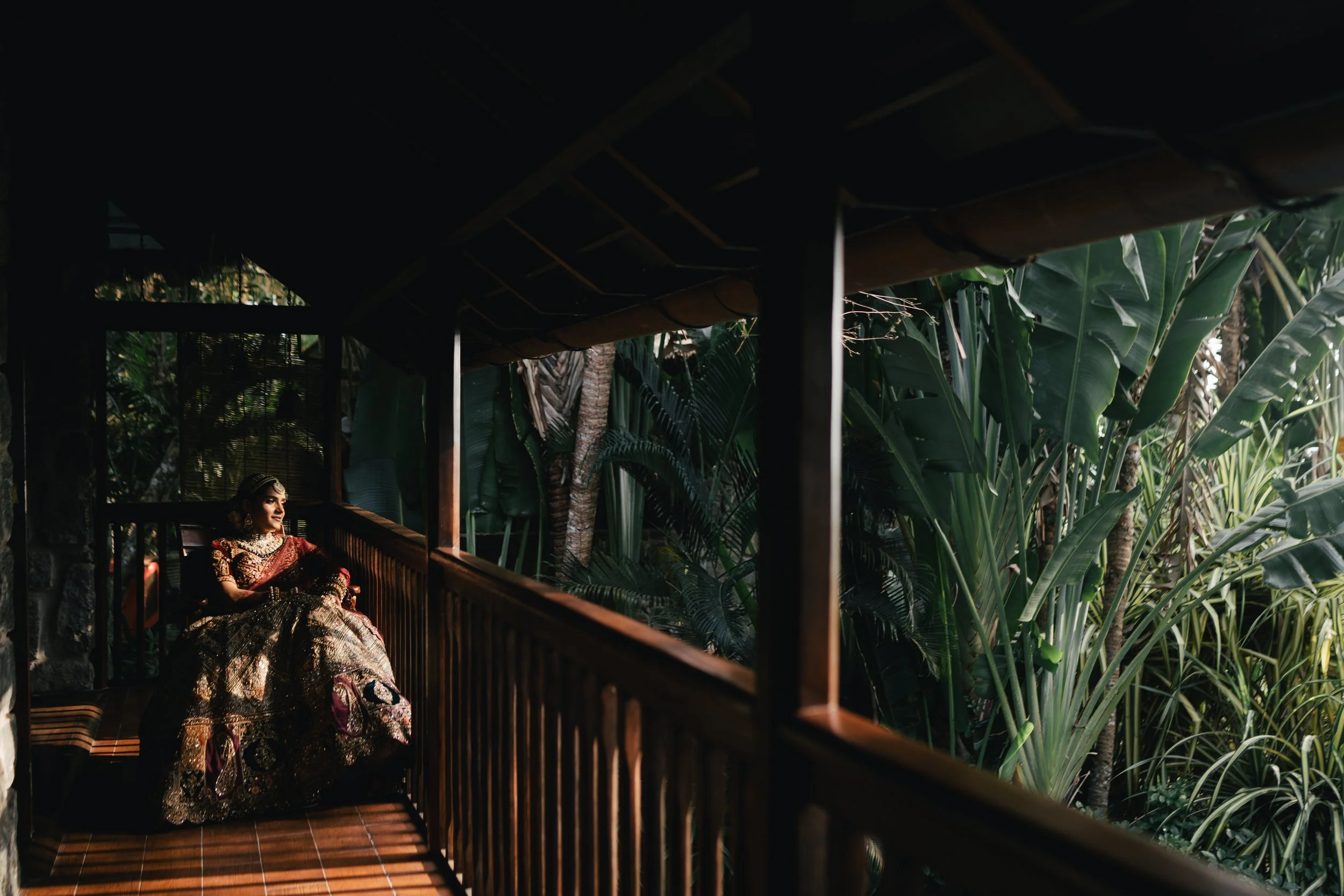 A woman dressed in traditional Indian attire, sitting on a balcony surrounded by lush tropical plants.