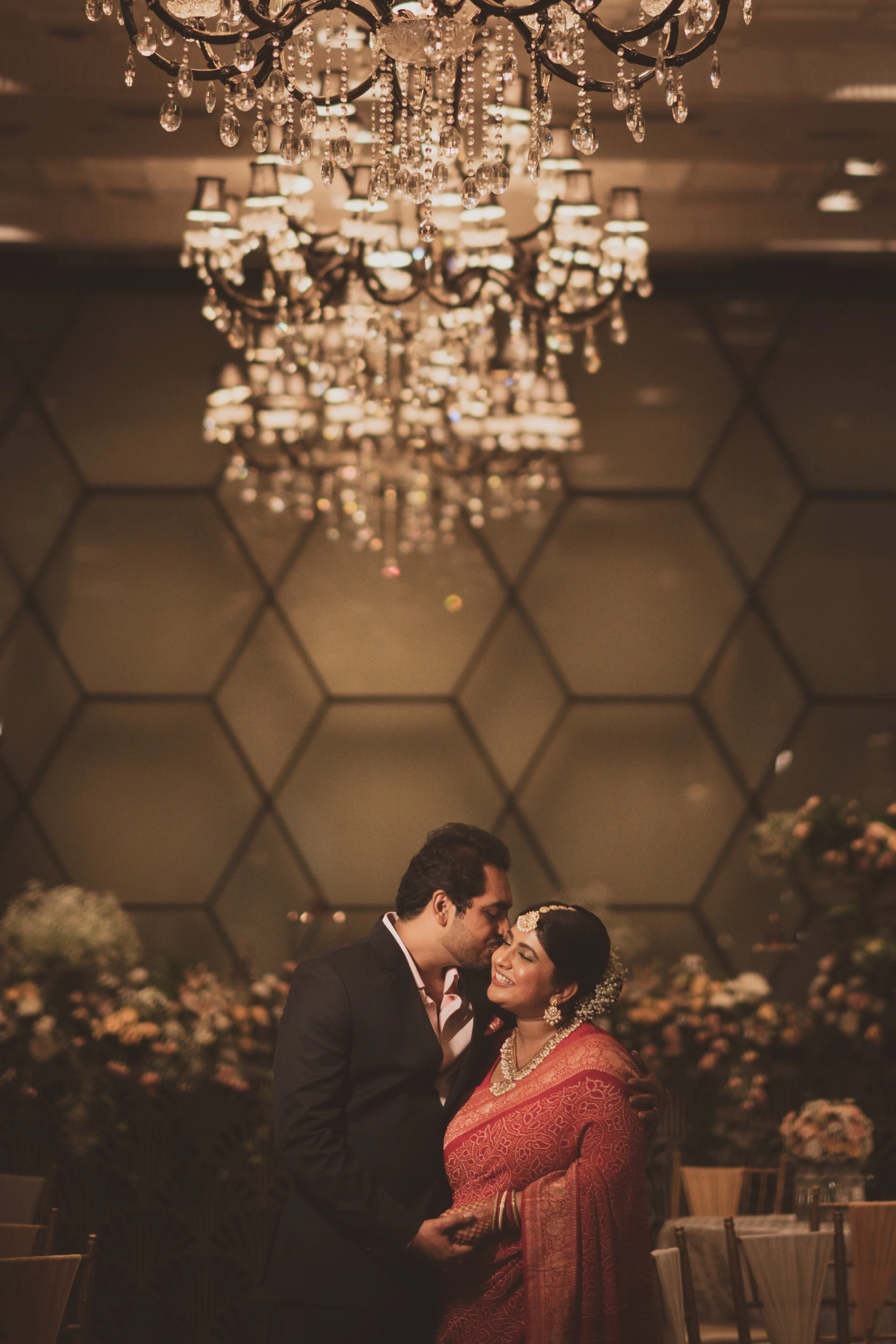 A couple dressed in formal attire embracing each other at a wedding reception, with chandeliers hanging from the ceiling and floral arrangements in the background.