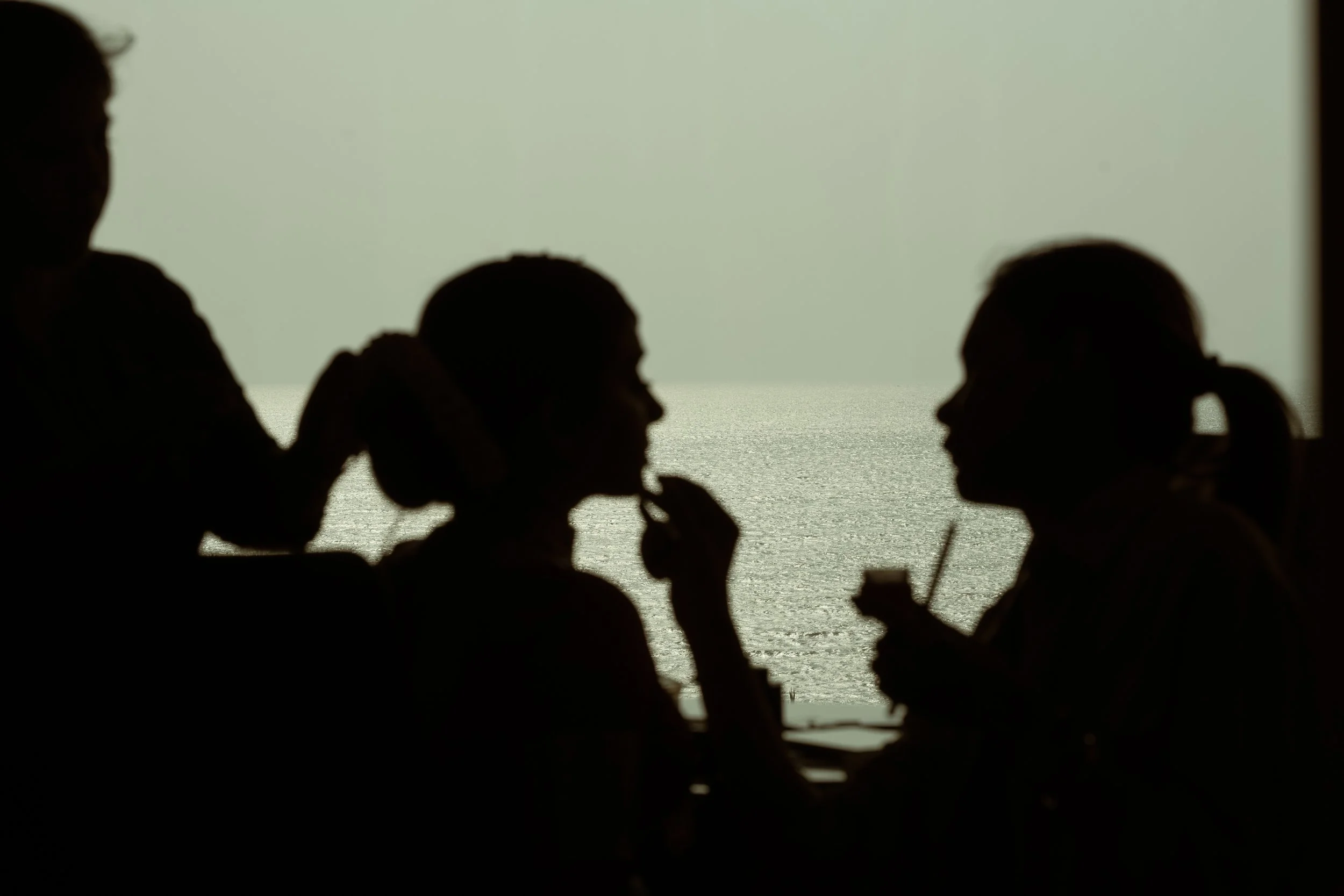 Silhouettes of four women in a restaurant or cafe, with one woman eating and others talking, with a view of the ocean in the background.