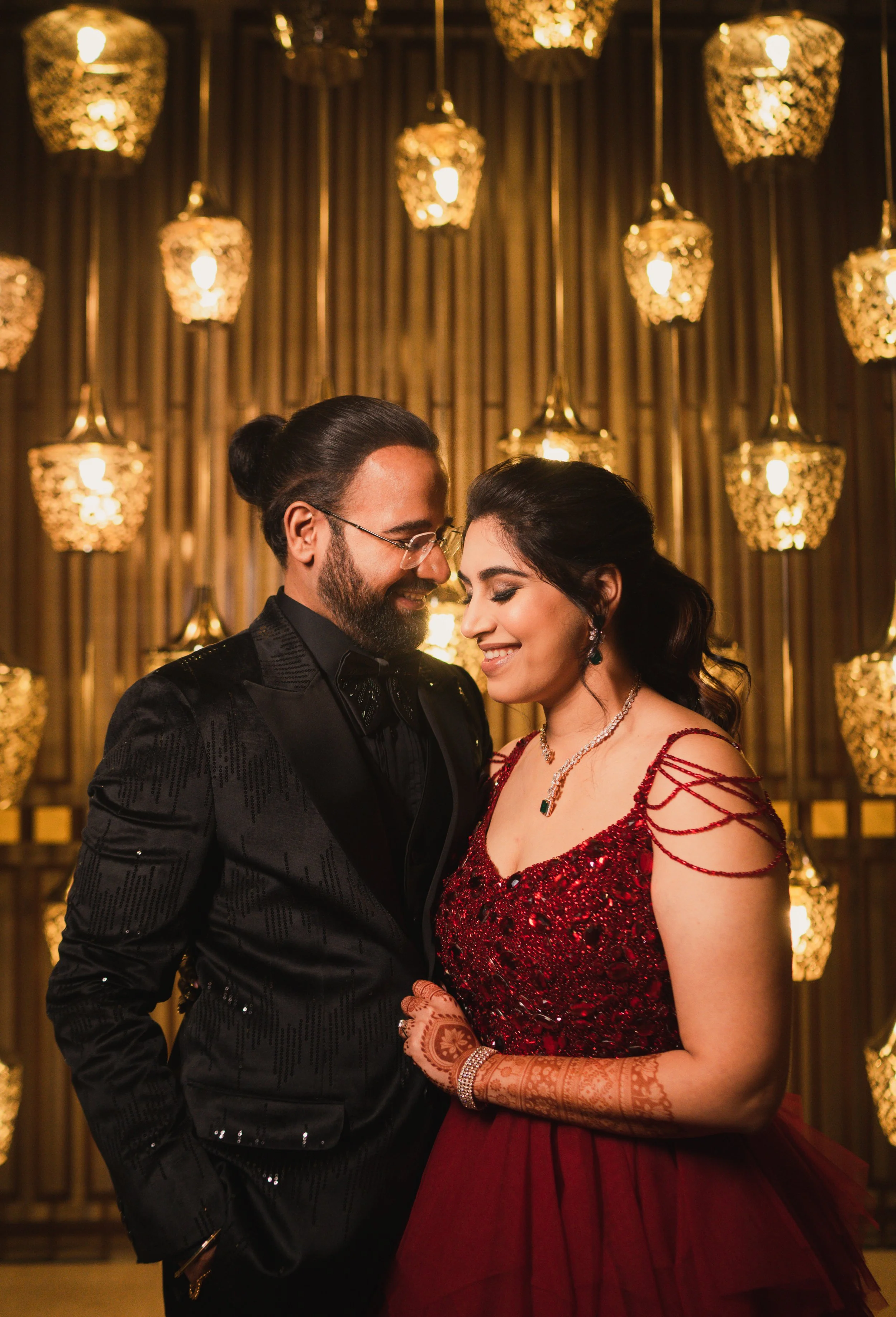 A couple dressed in elegant formal attire, smiling and close to each other at an event with warm decorative hanging lights in the background.