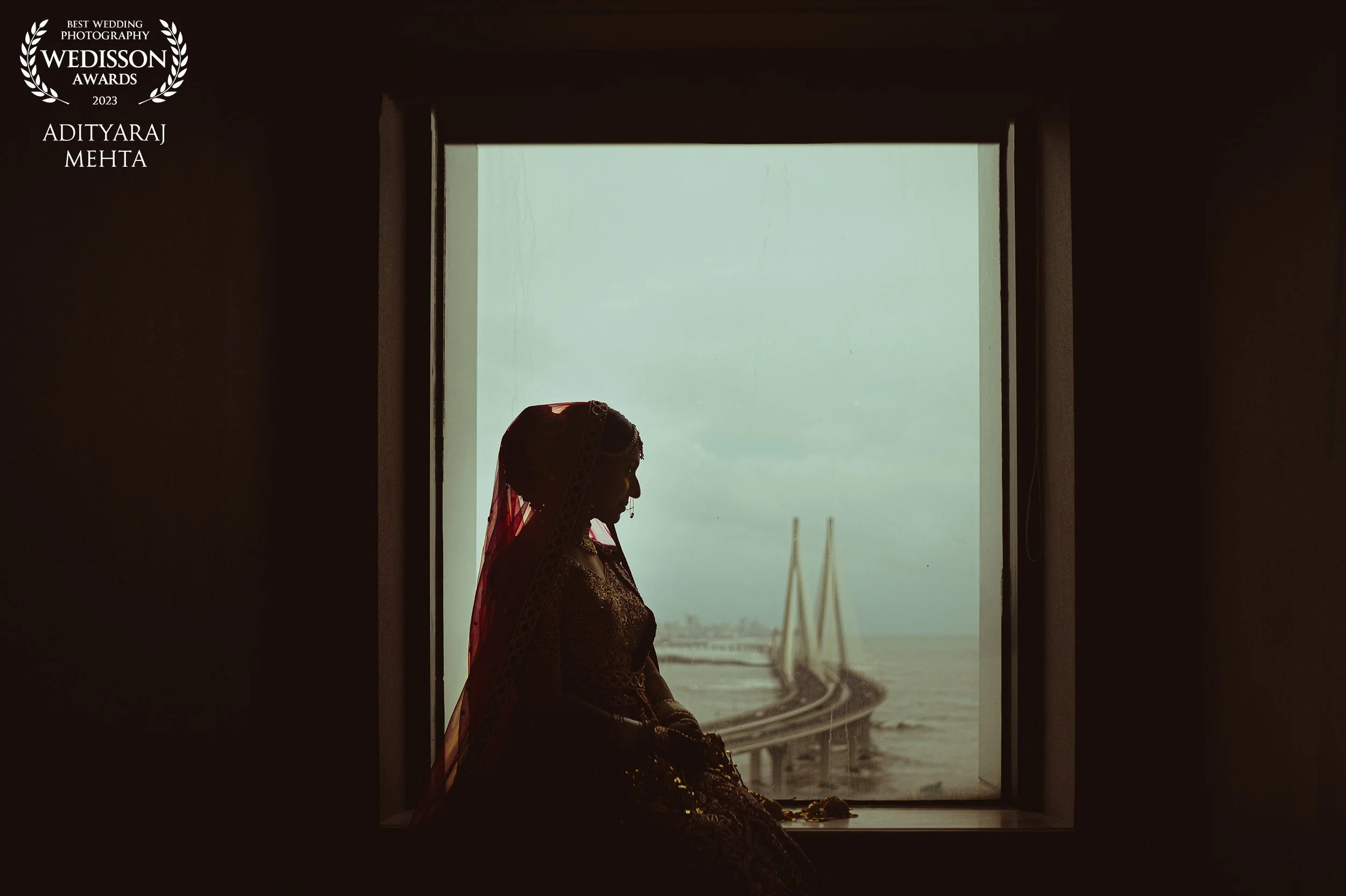 Silhouette of a woman in traditional attire sitting by a window with a bridge over water visible outside.