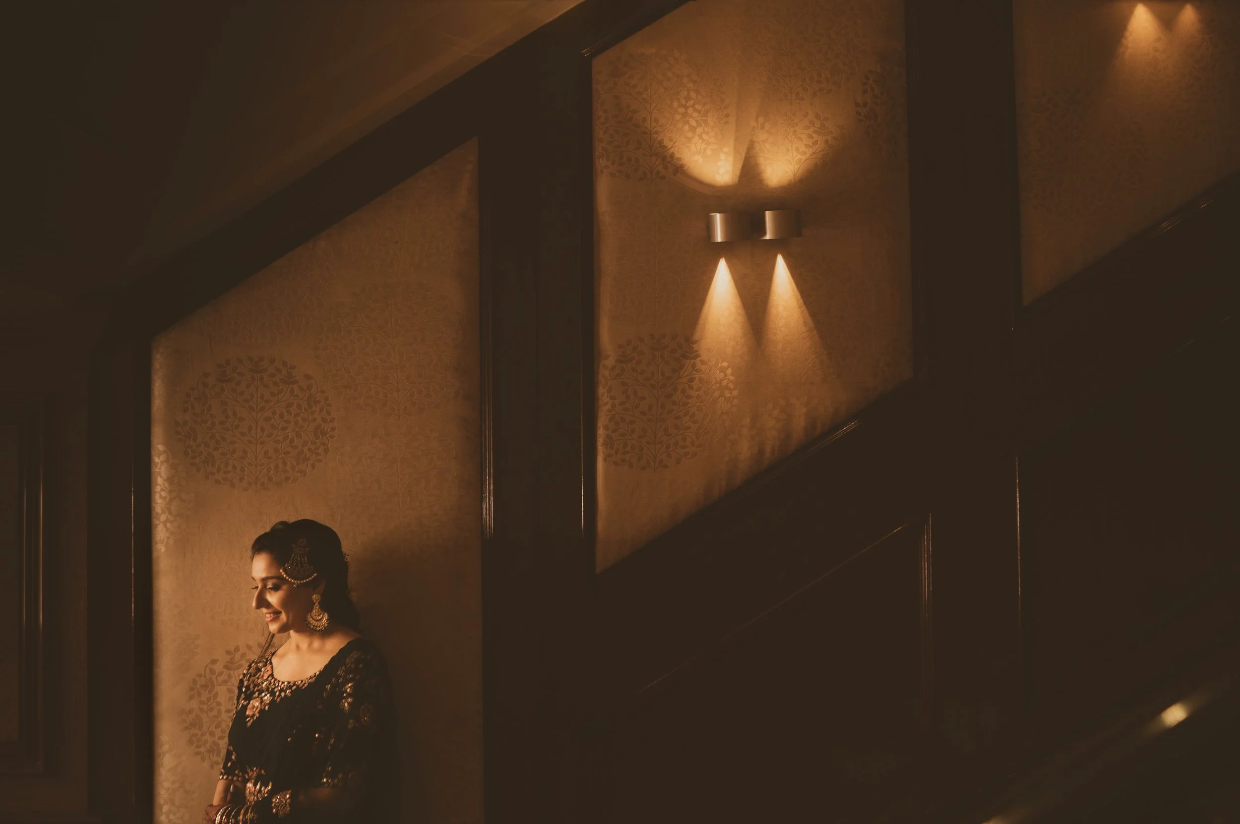 A woman dressed in traditional Indian attire, wearing jewelry, smiling and standing near a patterned wall illuminated by soft lighting.