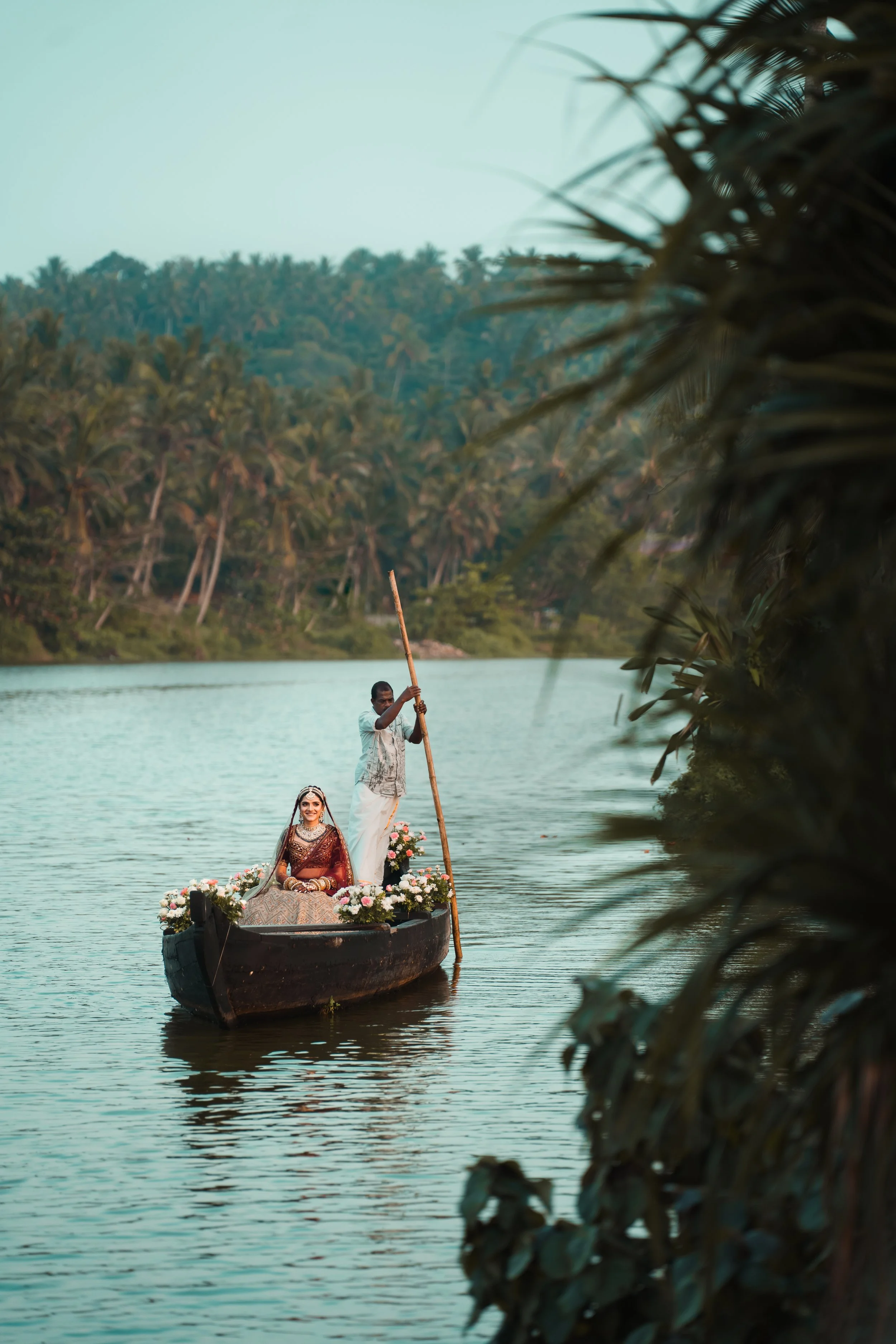 A woman dressed in traditional Indian clothing sitting on a boat decorated with flowers, being rowed by a man on a body of water with a lush, green, tropical forest background.