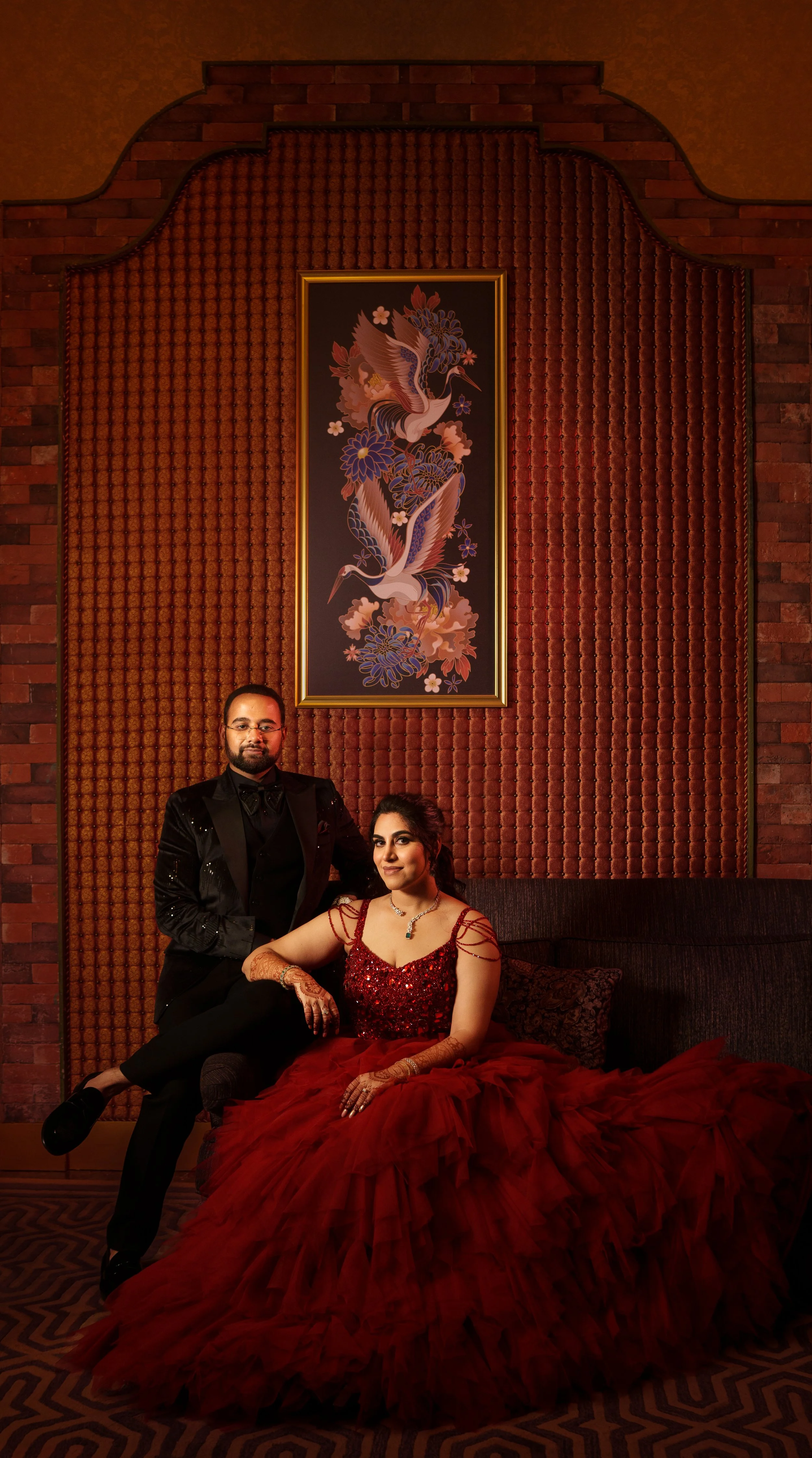 A man in a black tuxedo and a woman in a red gown with intricate henna designs on her hands pose together in a luxurious room with warm lighting, patterned carpet, a textured wall, and a framed artwork of cranes and flowers.