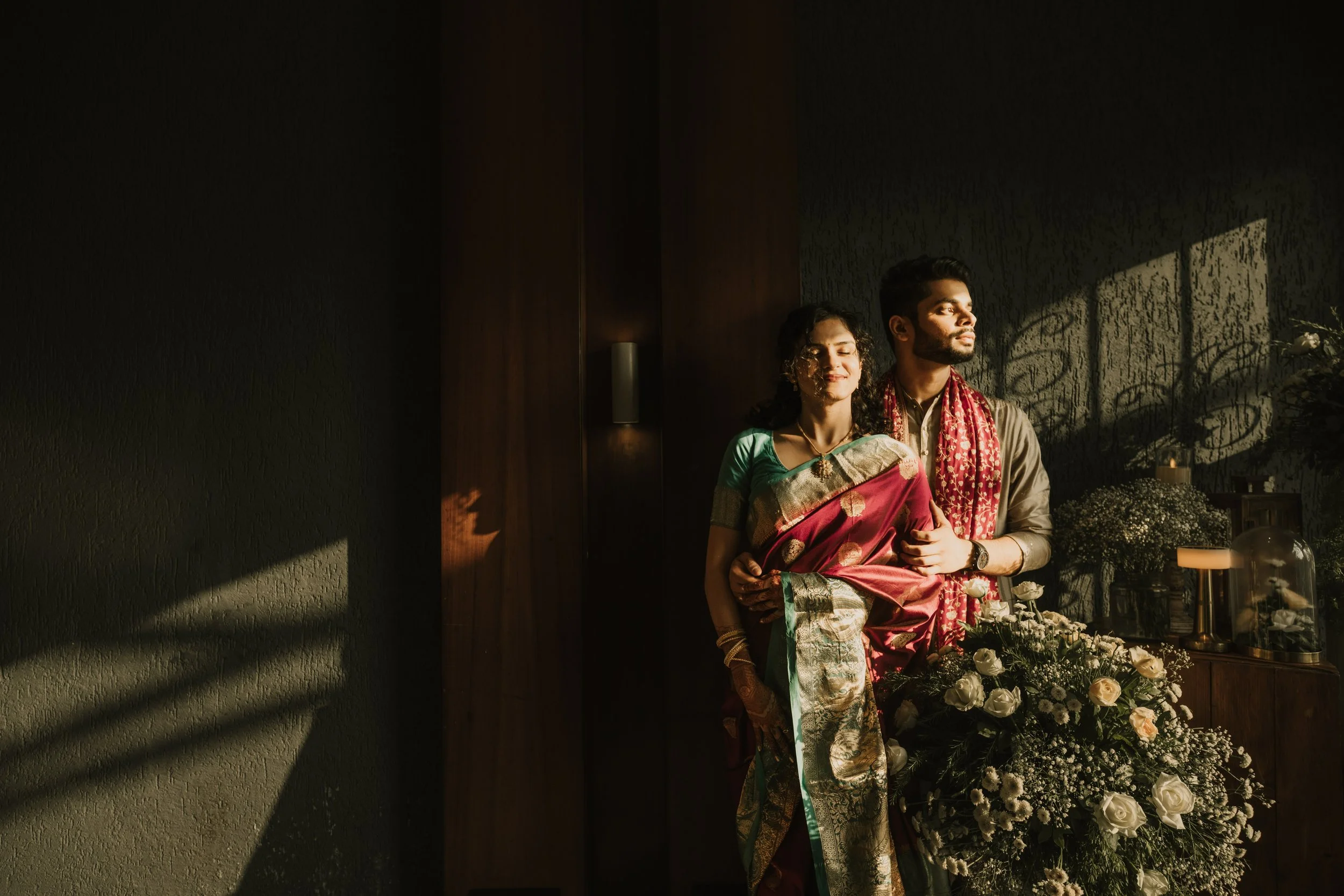 A couple stands together in warm sunlight, with a large floral arrangement in front of them, and a dark wall with subdued lighting in the background.