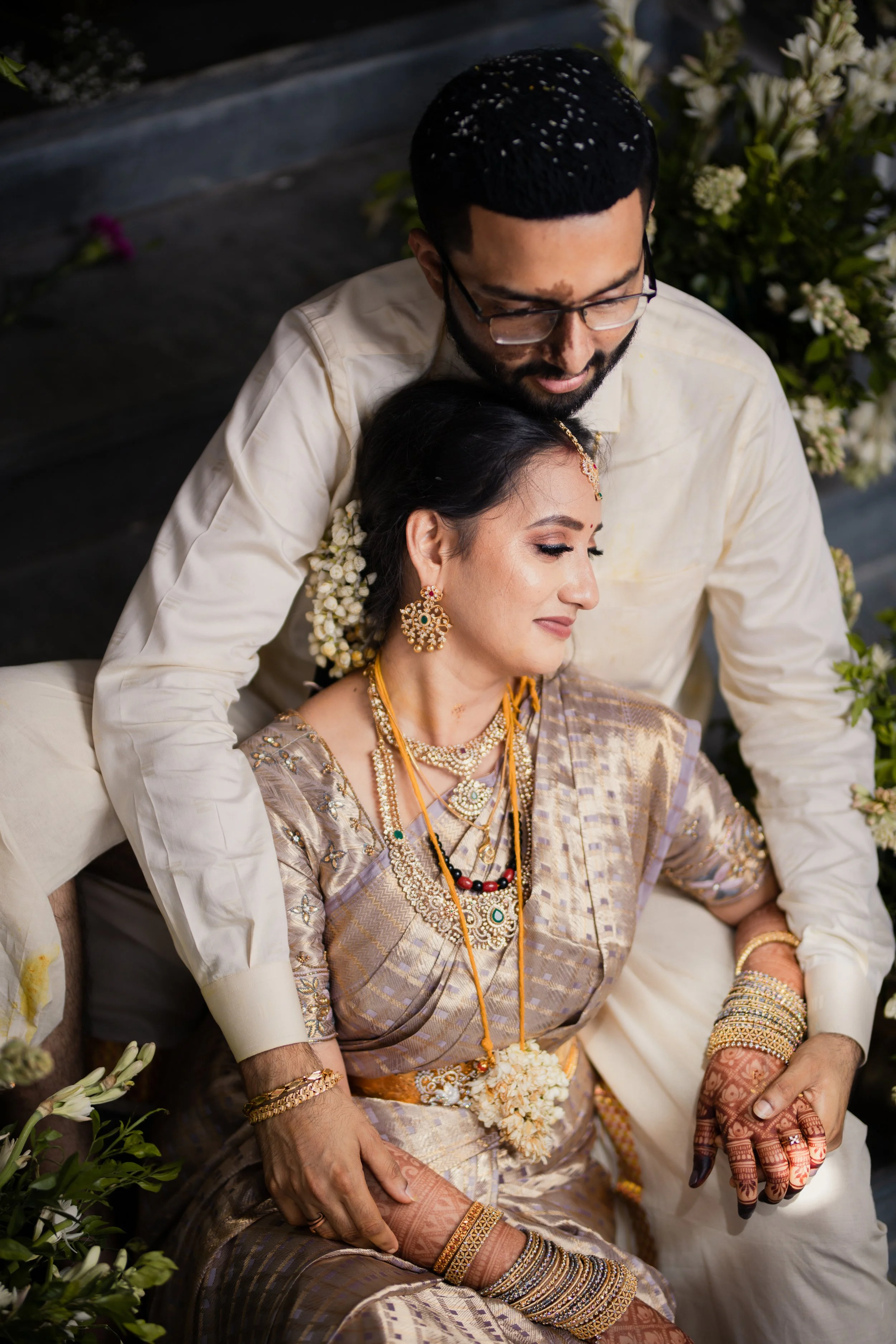 A bride and groom dressed in traditional Indian wedding attire, seated surrounded by flowers, with the groom standing behind the bride, both showing expressions of contentment and serenity.