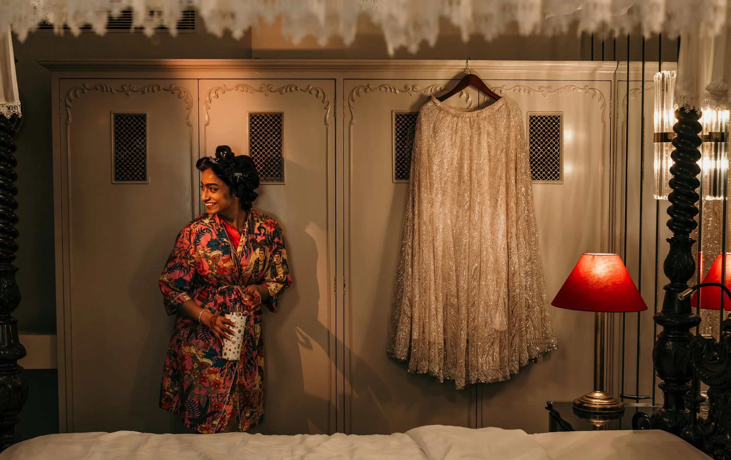Woman in a colorful robe standing in a room with a wardrobe, a gold dress hanging, and a red lamp on a bedside table.