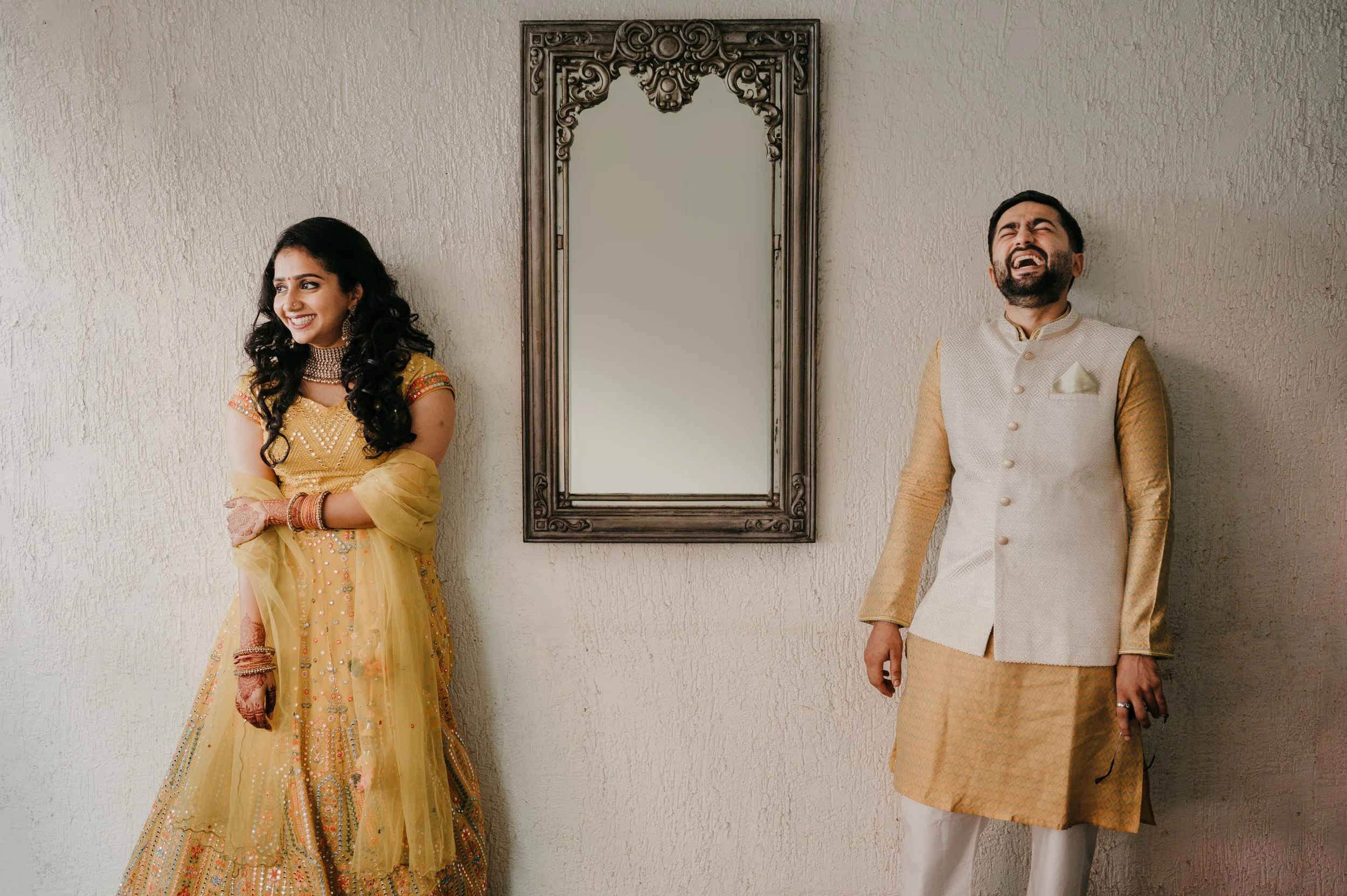 A man in a cream and gold traditional Indian outfit laughing next to a woman in yellow traditional Indian attire with detailed embroidery and jewelry, both standing against a beige textured wall with a decorative mirror in the center.