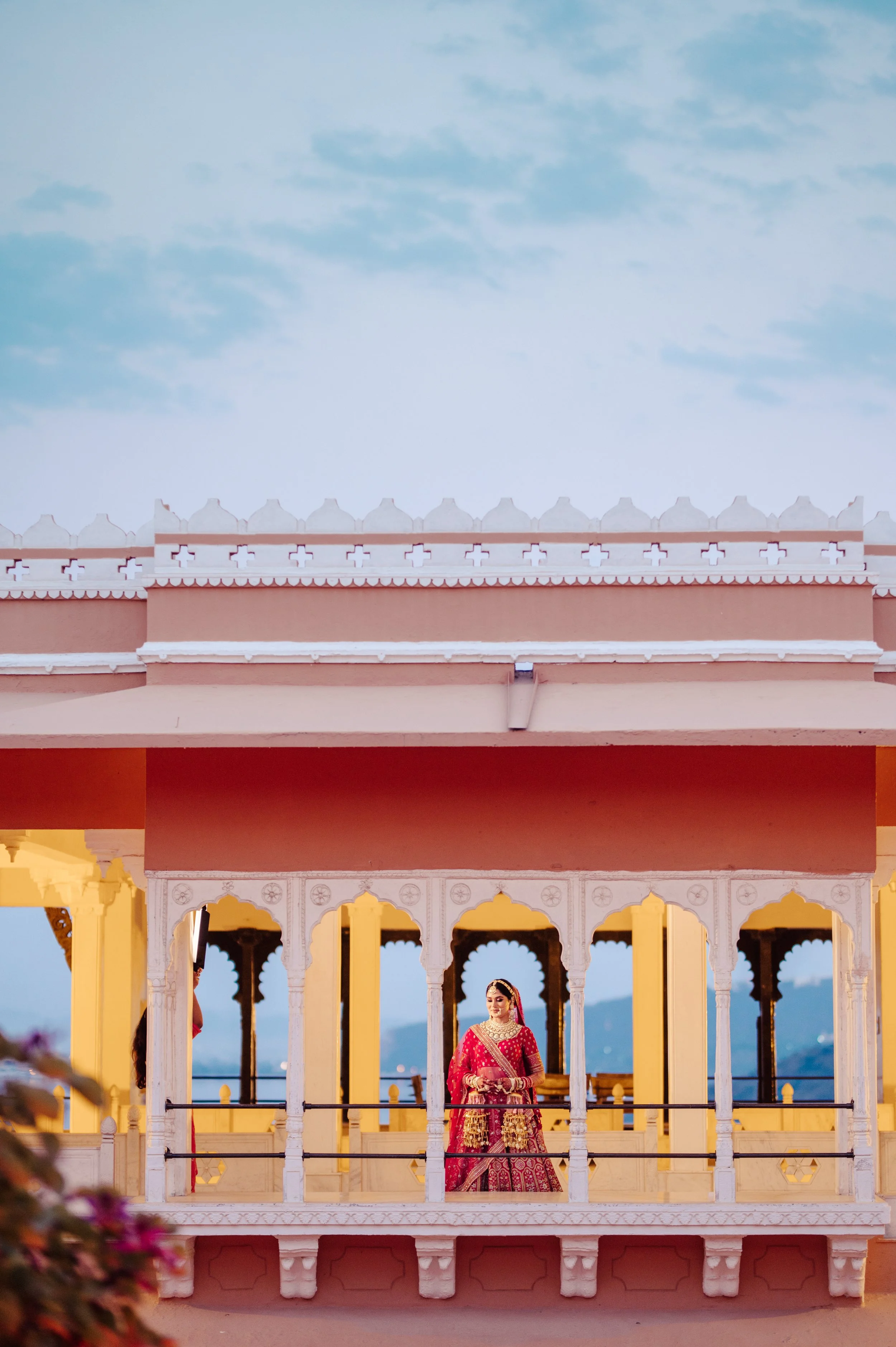 A woman in traditional Indian attire standing on a decorated balcony of a pink and yellow building with arches, against a backdrop of mountains and blue sky.