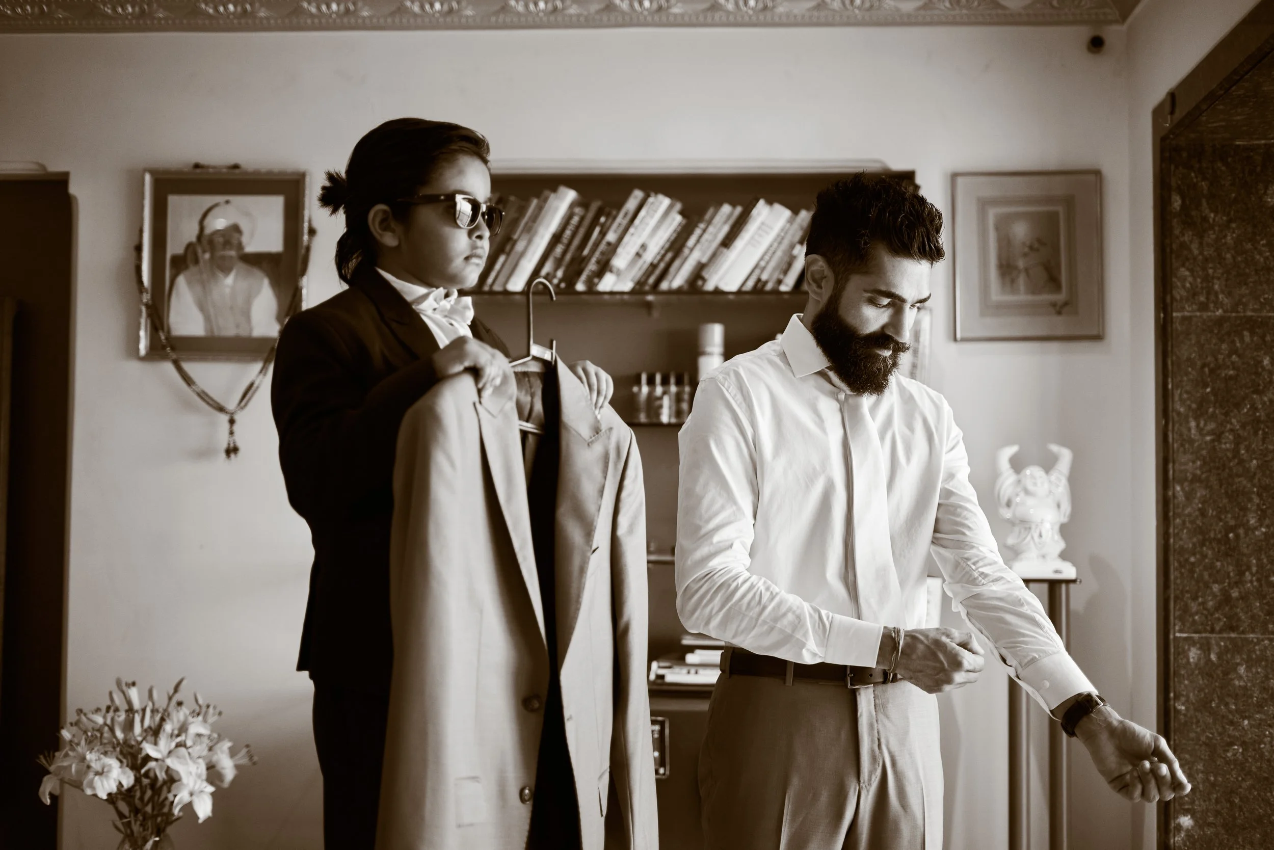 A man getting dressed while a woman holds a coat and hanger in a room with bookshelves and framed pictures.