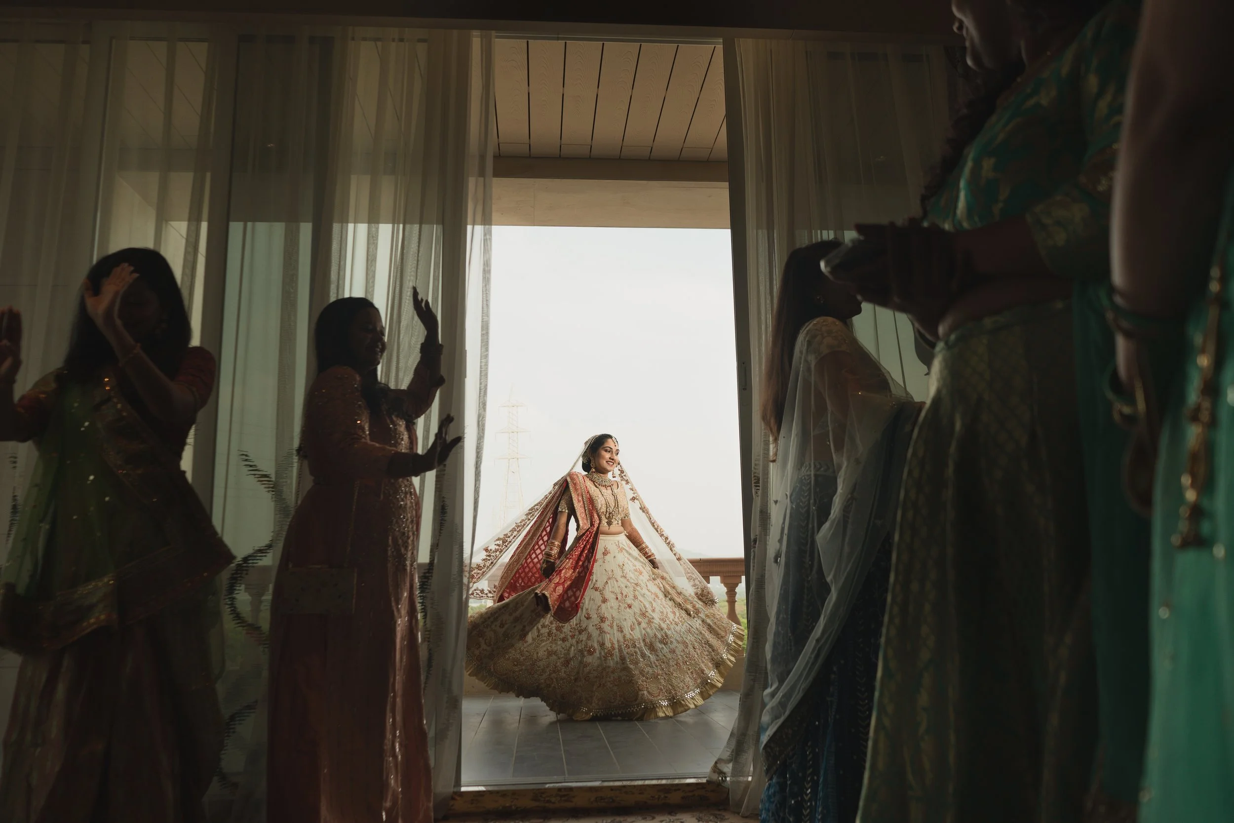 A bride in a traditional wedding dress standing on a balcony, framed by women dressed in colorful sarees on either side, during a pre-wedding celebration.