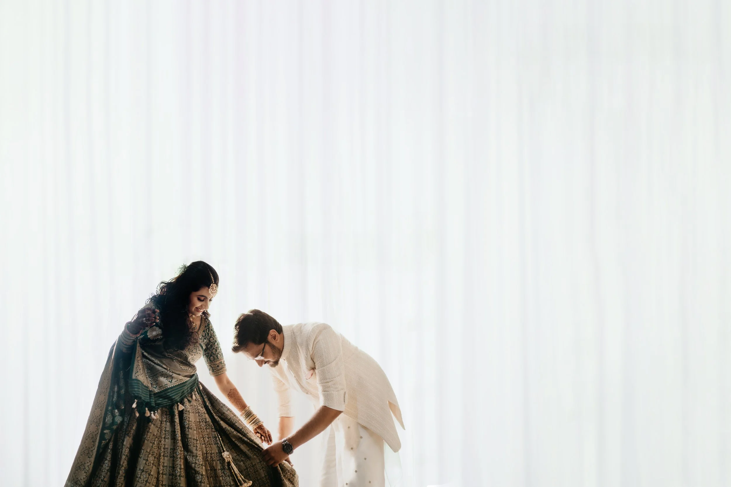 A couple dressed in traditional Indian wedding attire, with the woman wearing a lehenga and the man in a cream-colored sherwani, looking at the woman's dress while standing against a white background.