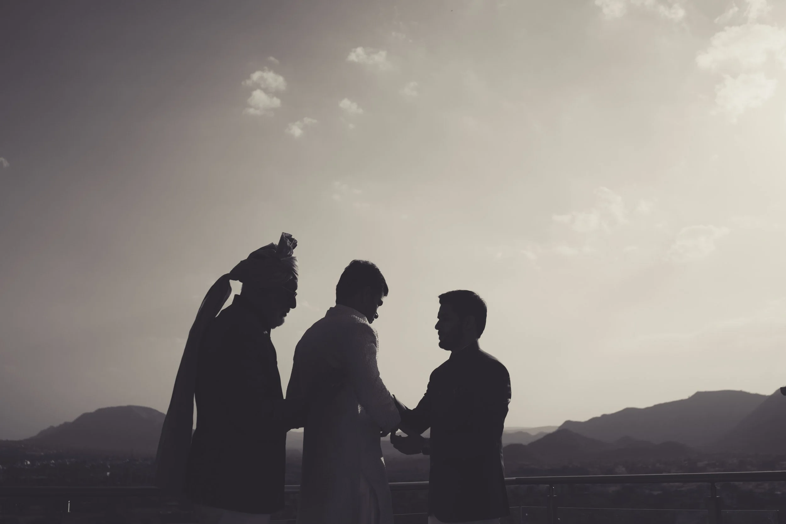 Silhouettes of three people shaking hands outdoors with mountains and cloudy sky in the background