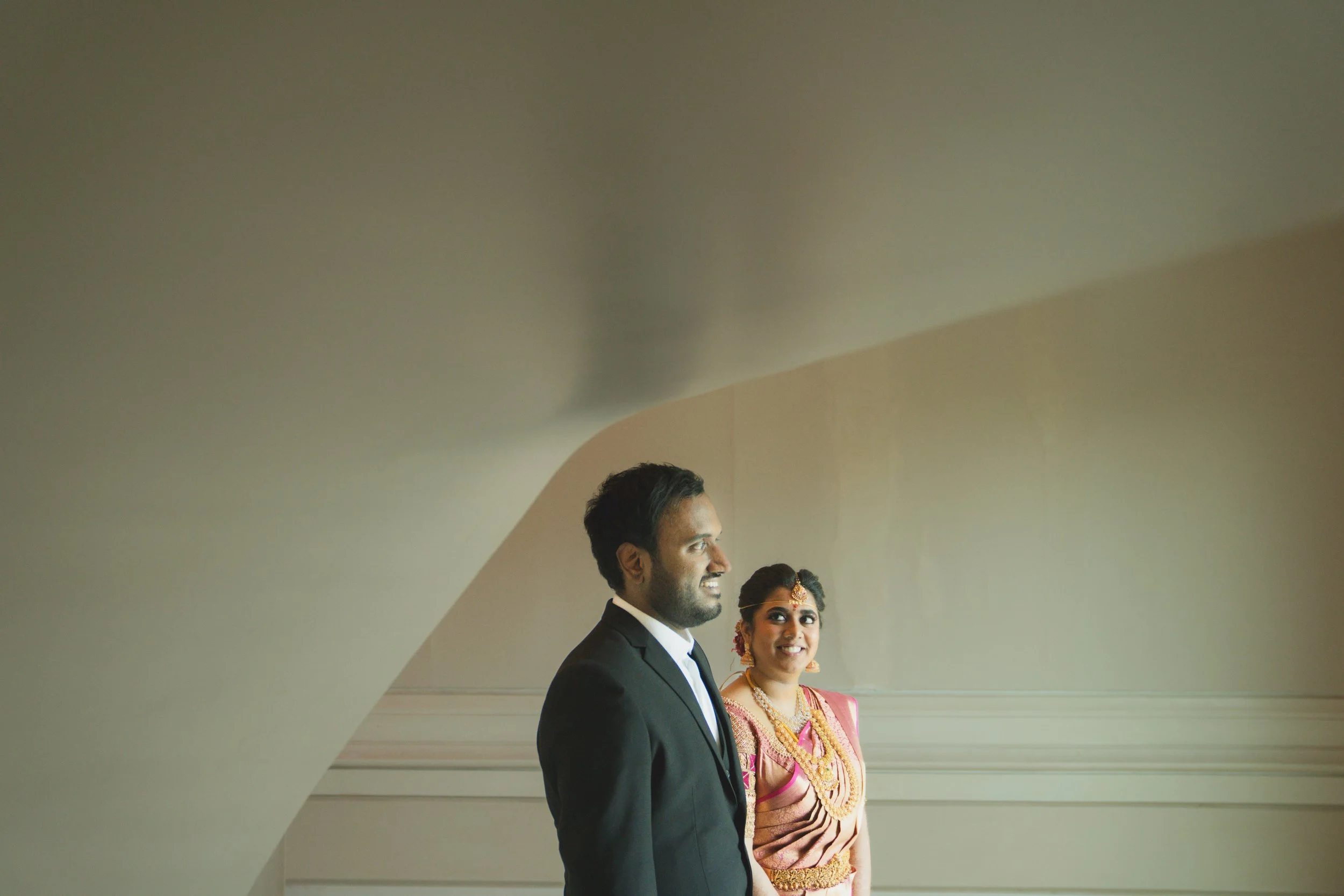 A groom in a black suit and a bride in traditional Indian attire standing inside a room with plain walls, with a large white curved ceiling above them.