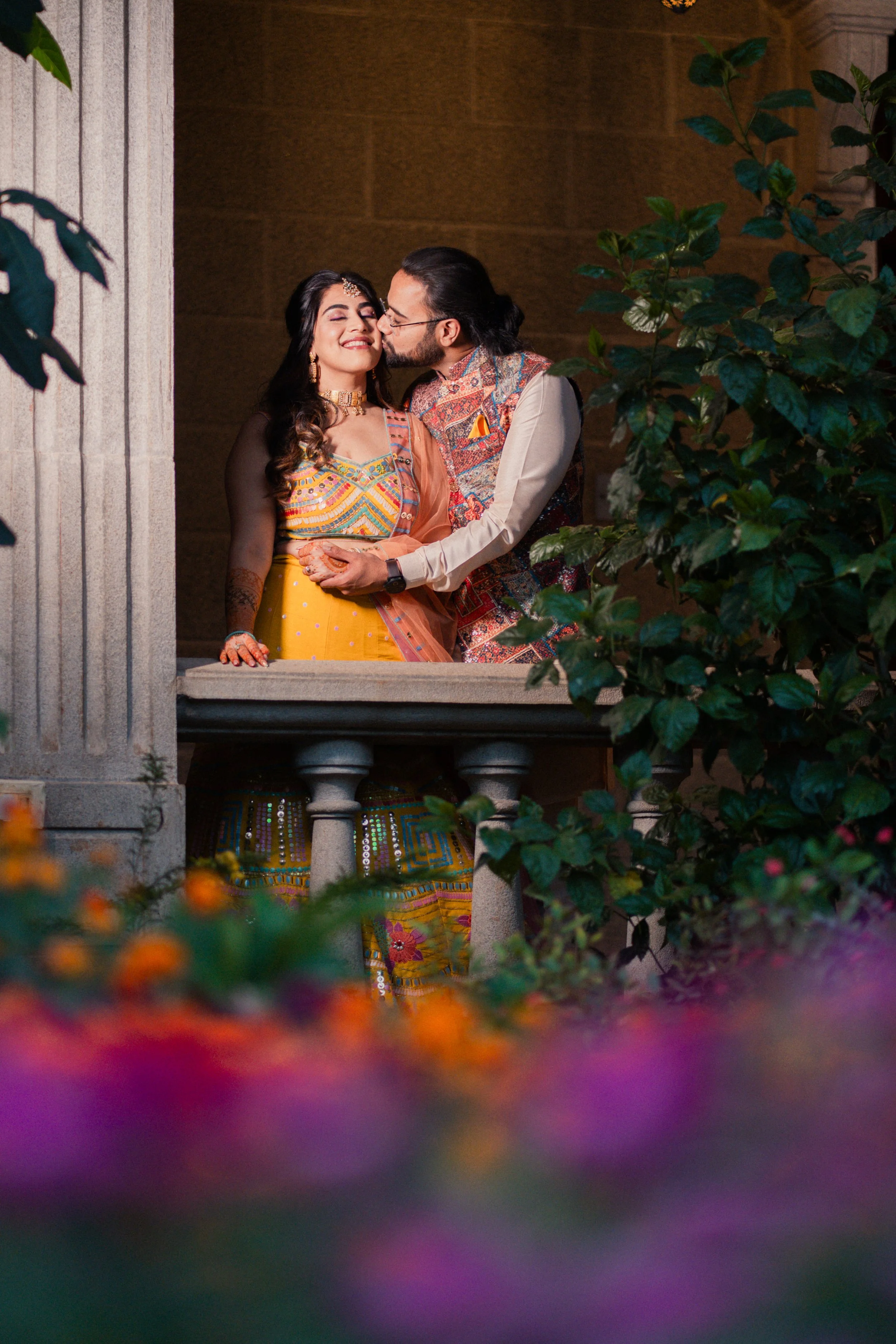 A couple dressed in traditional Indian attire sharing a romantic moment on a balcony surrounded by greenery and flowers.