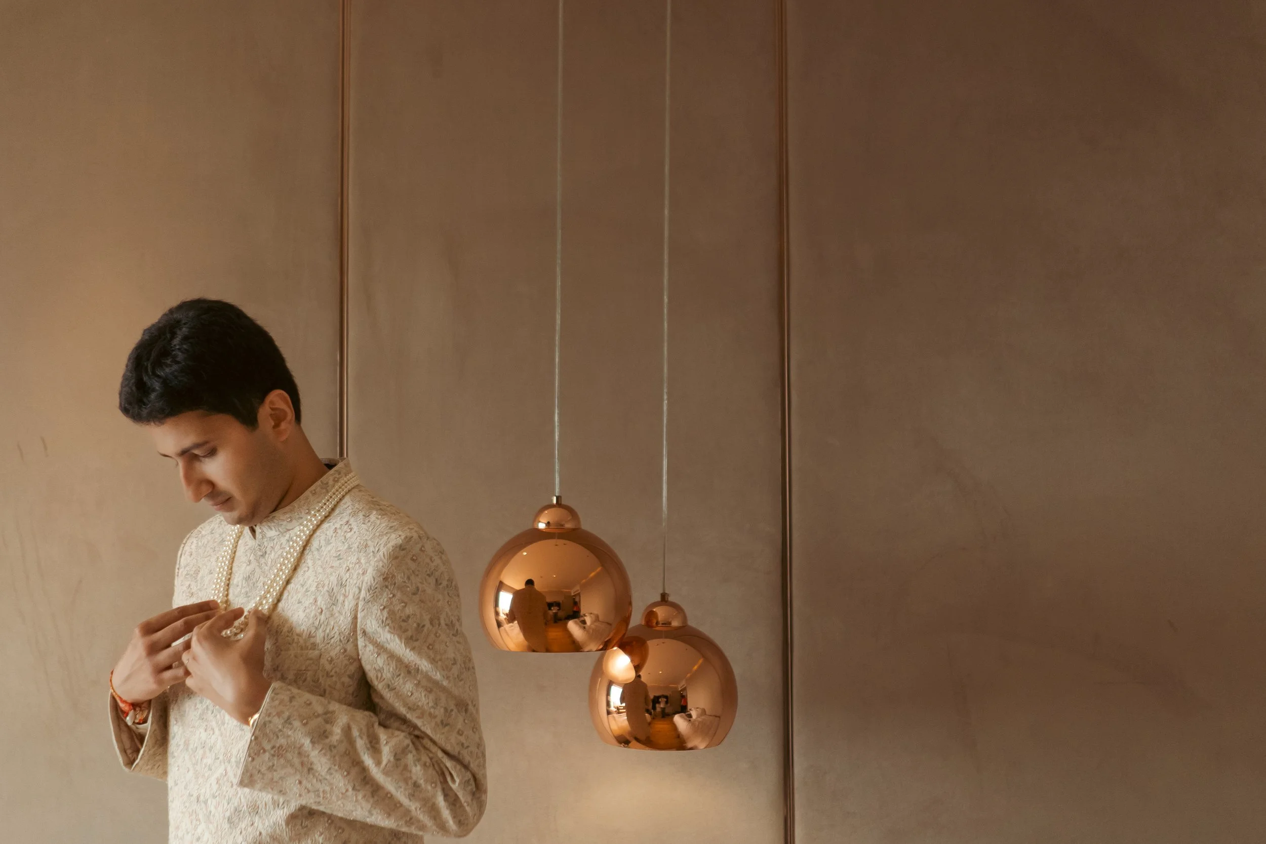 Young man wearing an ornate traditional Indian outfit, adjusting a gold necklace in a modern, minimalist interior with three hanging copper pendant lights.