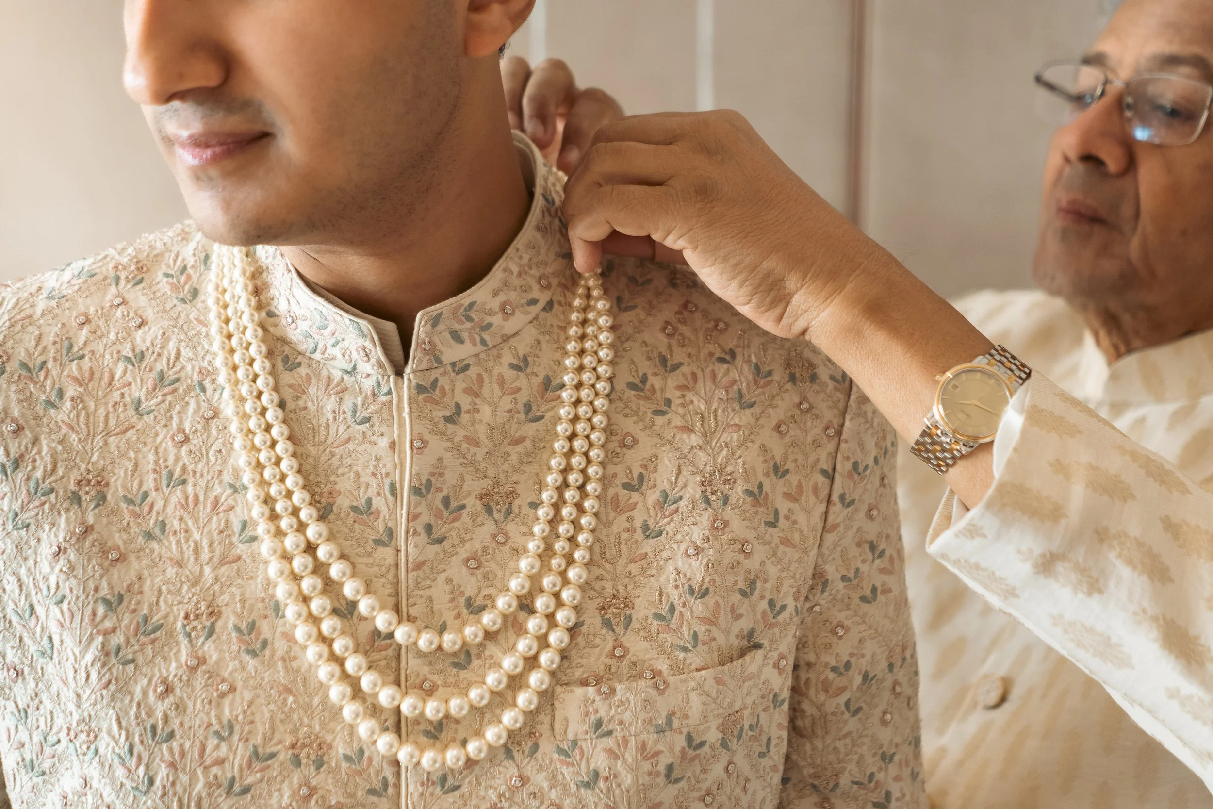 A man dressed in traditional cream-colored Indian attire with green and pink floral embroidery is being adorned with a pearl necklace by another man, who is also dressed in cream-colored traditional attire, while wearing a gold watch on his left wris