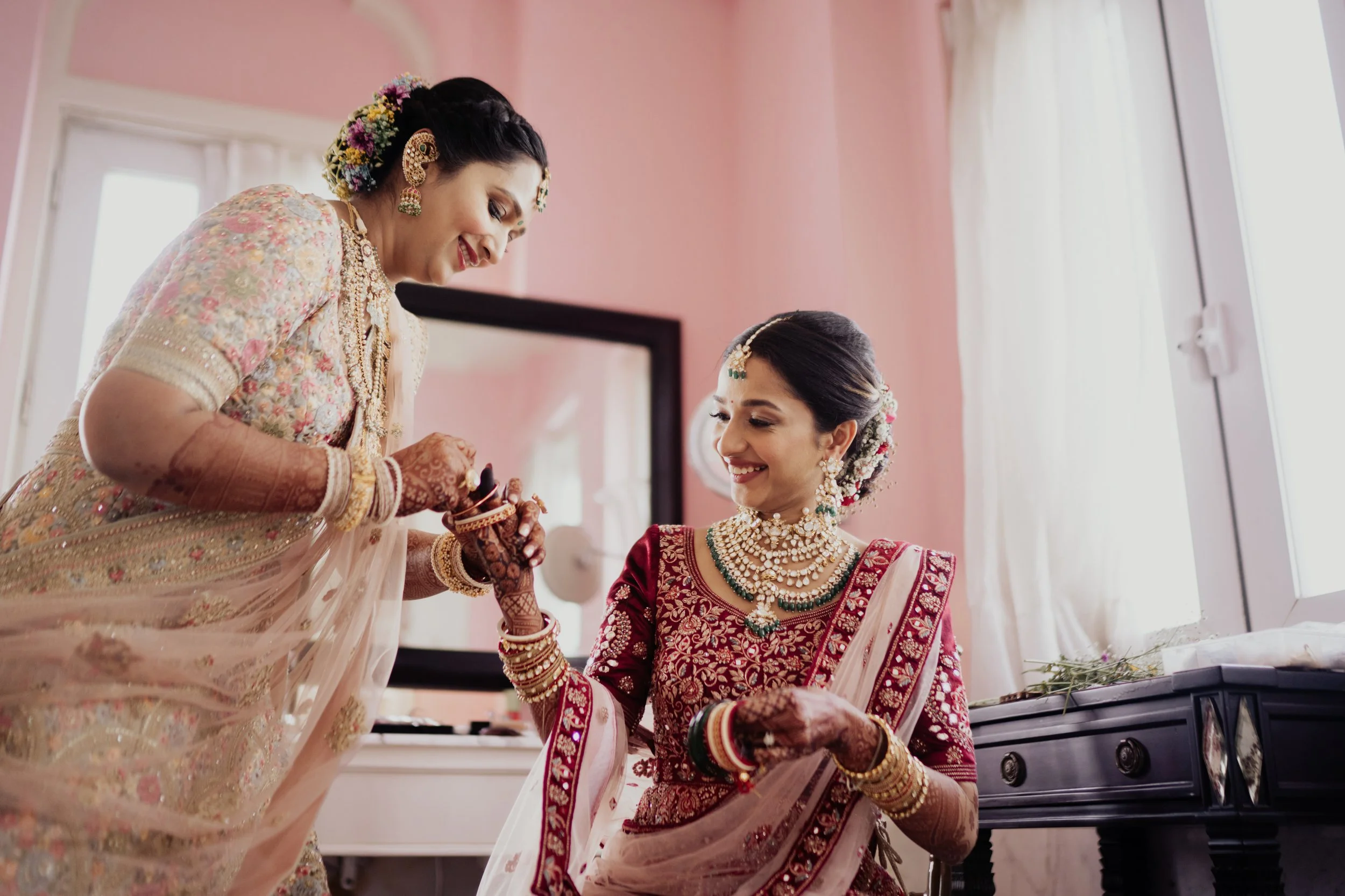 Two women dressed in traditional Indian wedding attire, with intricate jewelry, in a softly lit room with pink walls. One woman is standing, smiling, while the other is seated, also smiling, and holding the standing woman's hand.