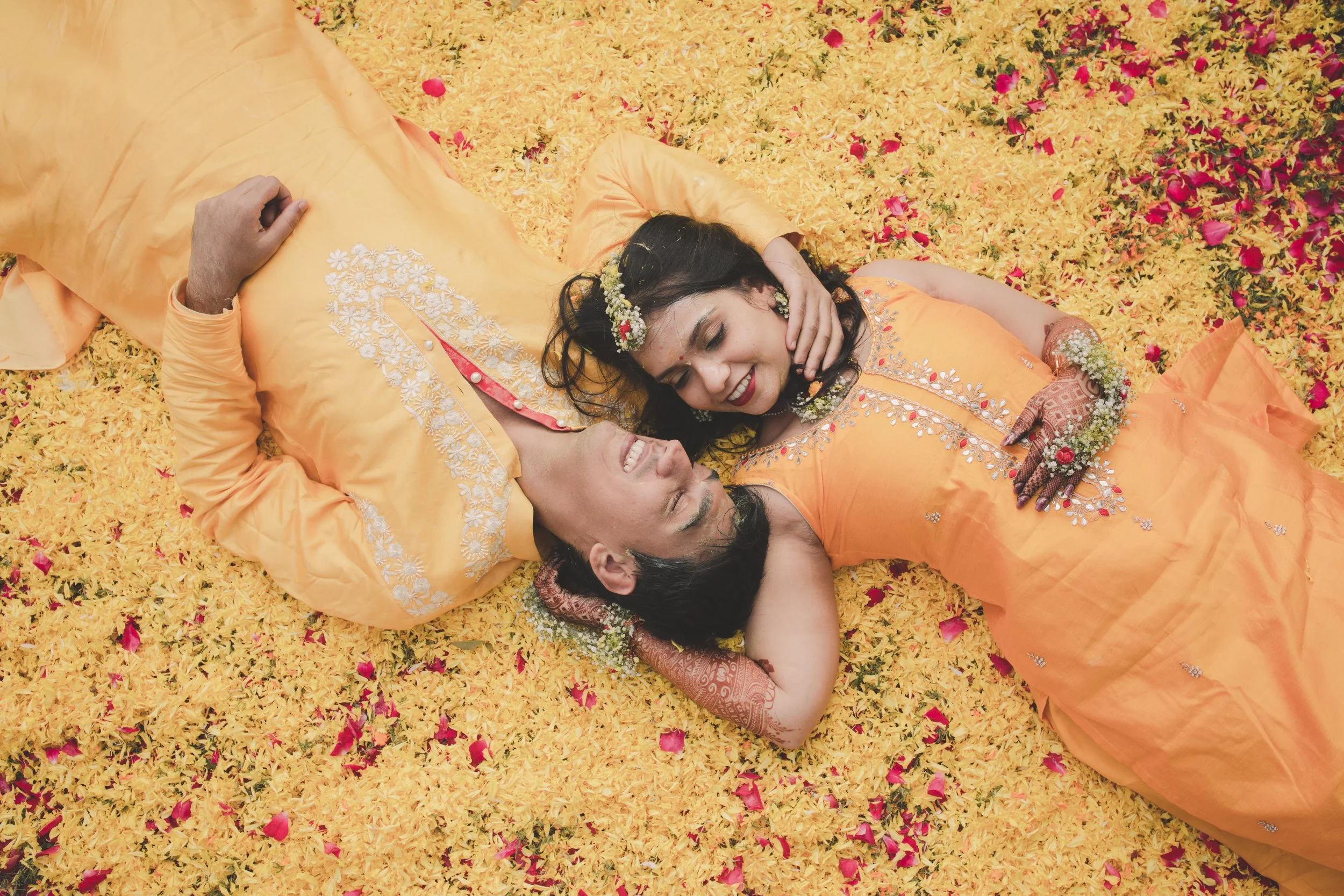 A couple dressed in traditional orange and yellow attire lying on a bed of yellow flower petals, smiling and looking at each other, surrounded by pink flower petals.