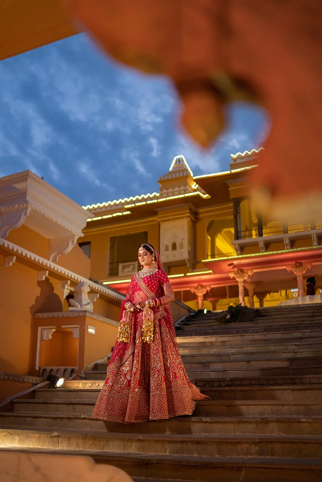 A woman in a traditional red and gold Indian wedding dress stands on the stairs outside a lit building at dusk, with a partly cloudy sky overhead.