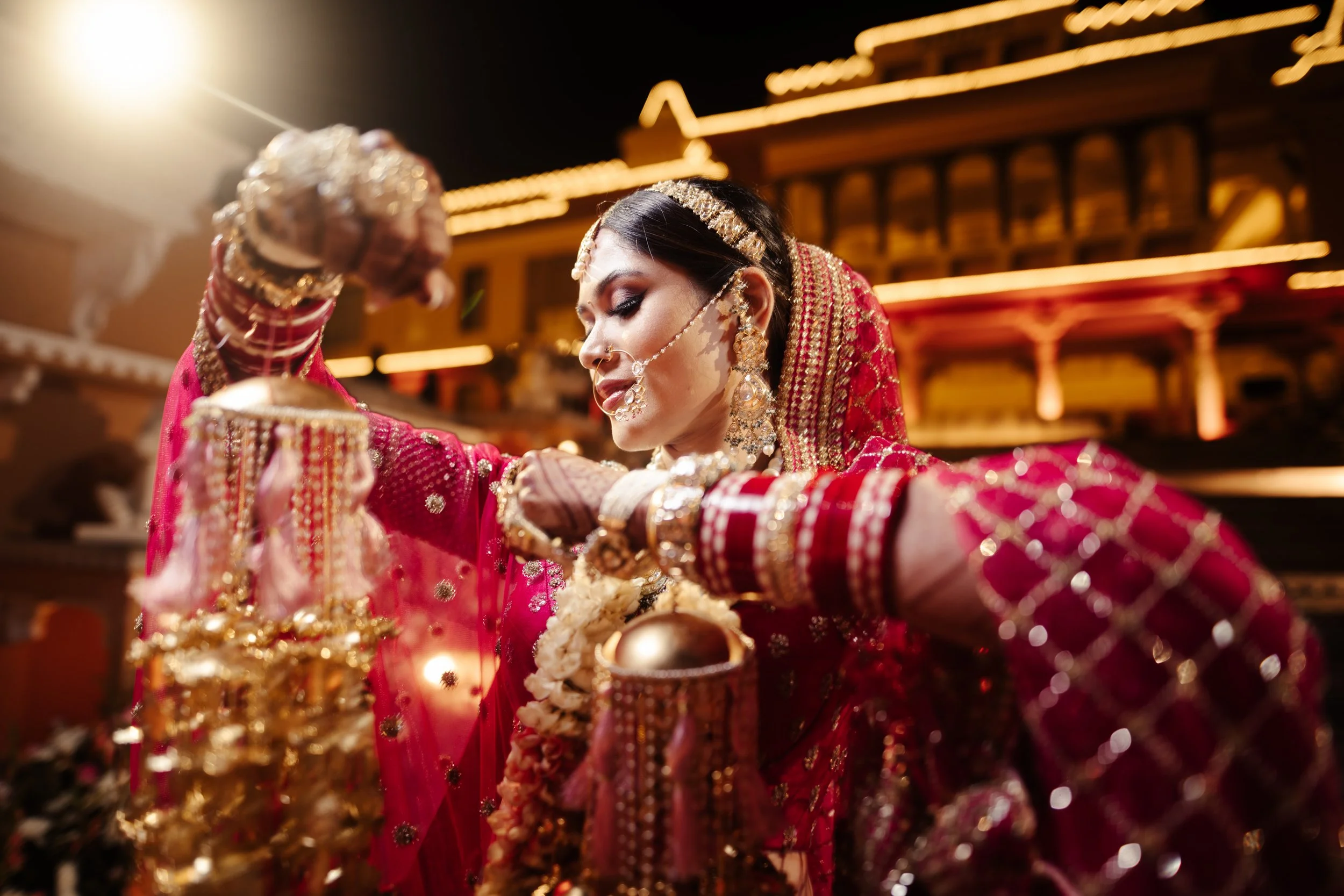 A bride dressed in red traditional Indian wedding attire and jewelry, performing a ritual at her wedding ceremony during night time.