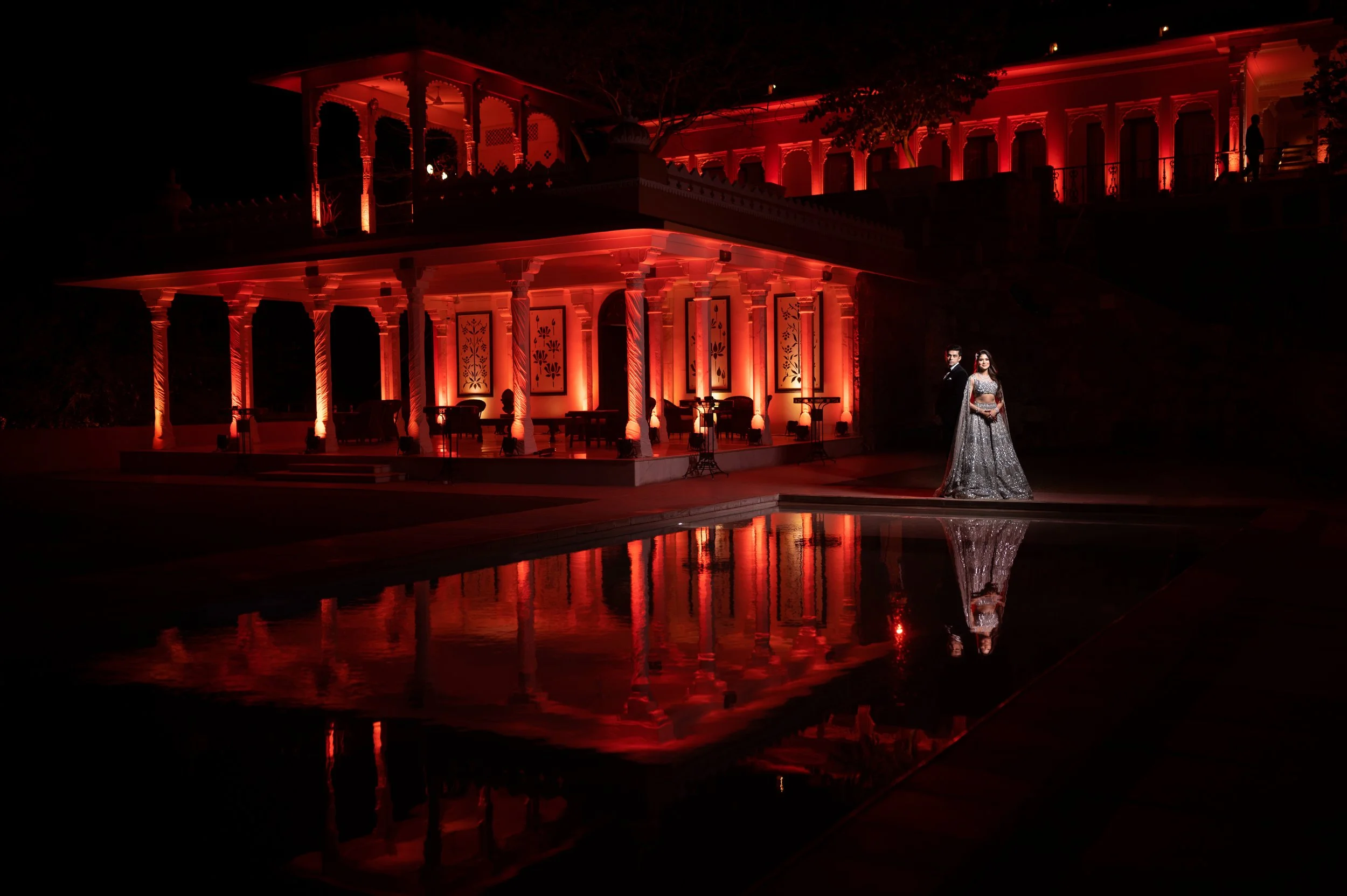A couple dressed in formal attire stands near a reflecting pool at night in front of a grand, multi-tiered building illuminated with red lights, with ornate architecture and trees around.