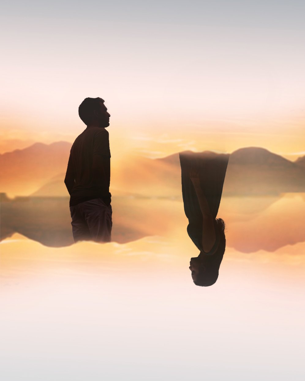 Silhouette of a man standing and a woman sitting reversed, against a mountain landscape at sunset, with their reflections mirrored on water.