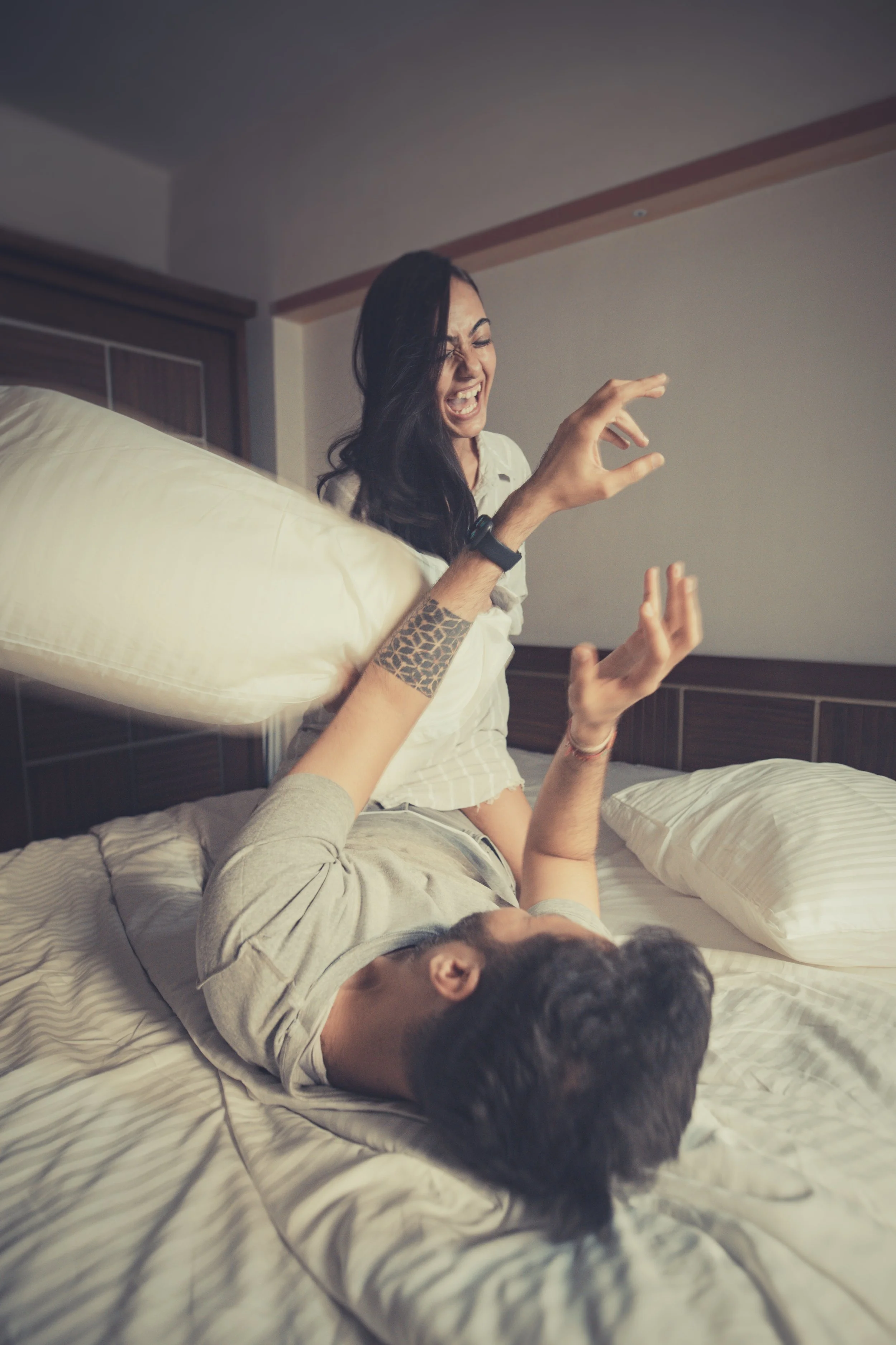 A woman sitting on top of a man lying on a bed, engaging in playful pillow fight with both smiling and laughing.