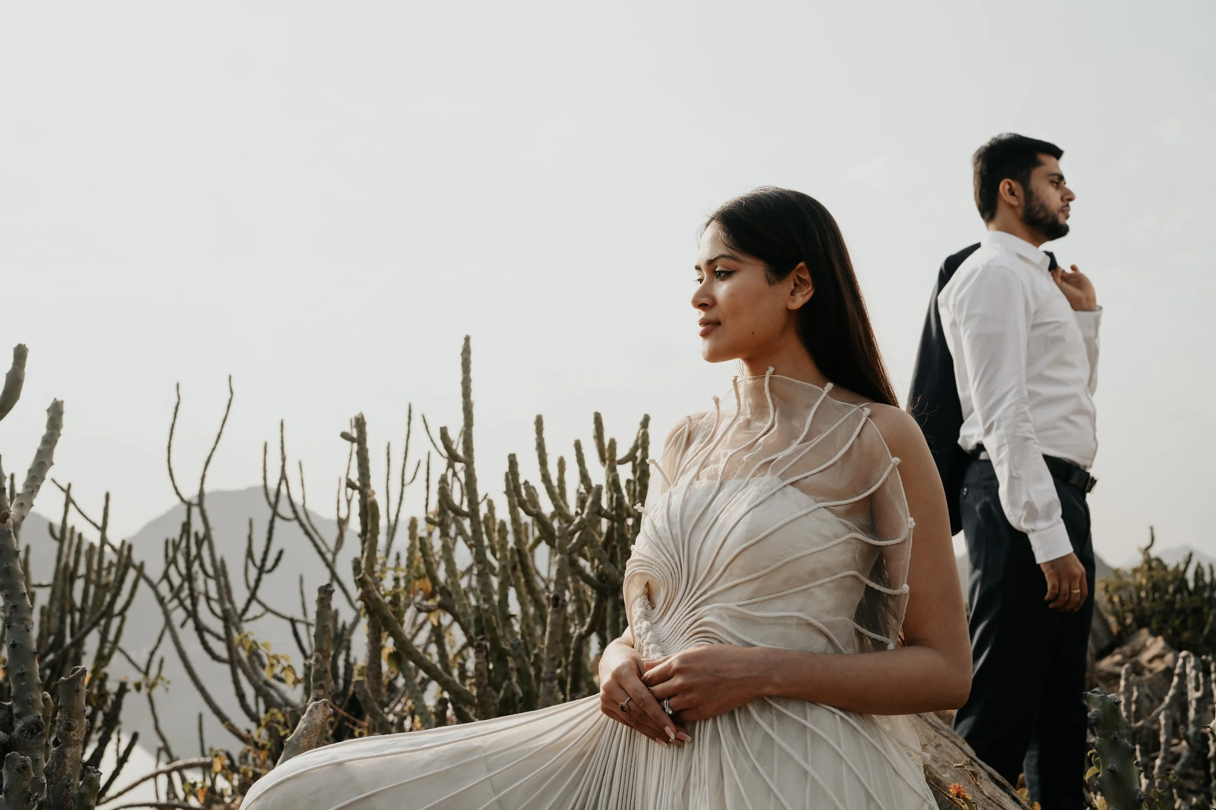 A woman in an elegant dress sits on a rock in a desert landscape with cacti, while a man in a white shirt and black pants stands nearby with a jacket over his shoulder, against a cloudy sky.