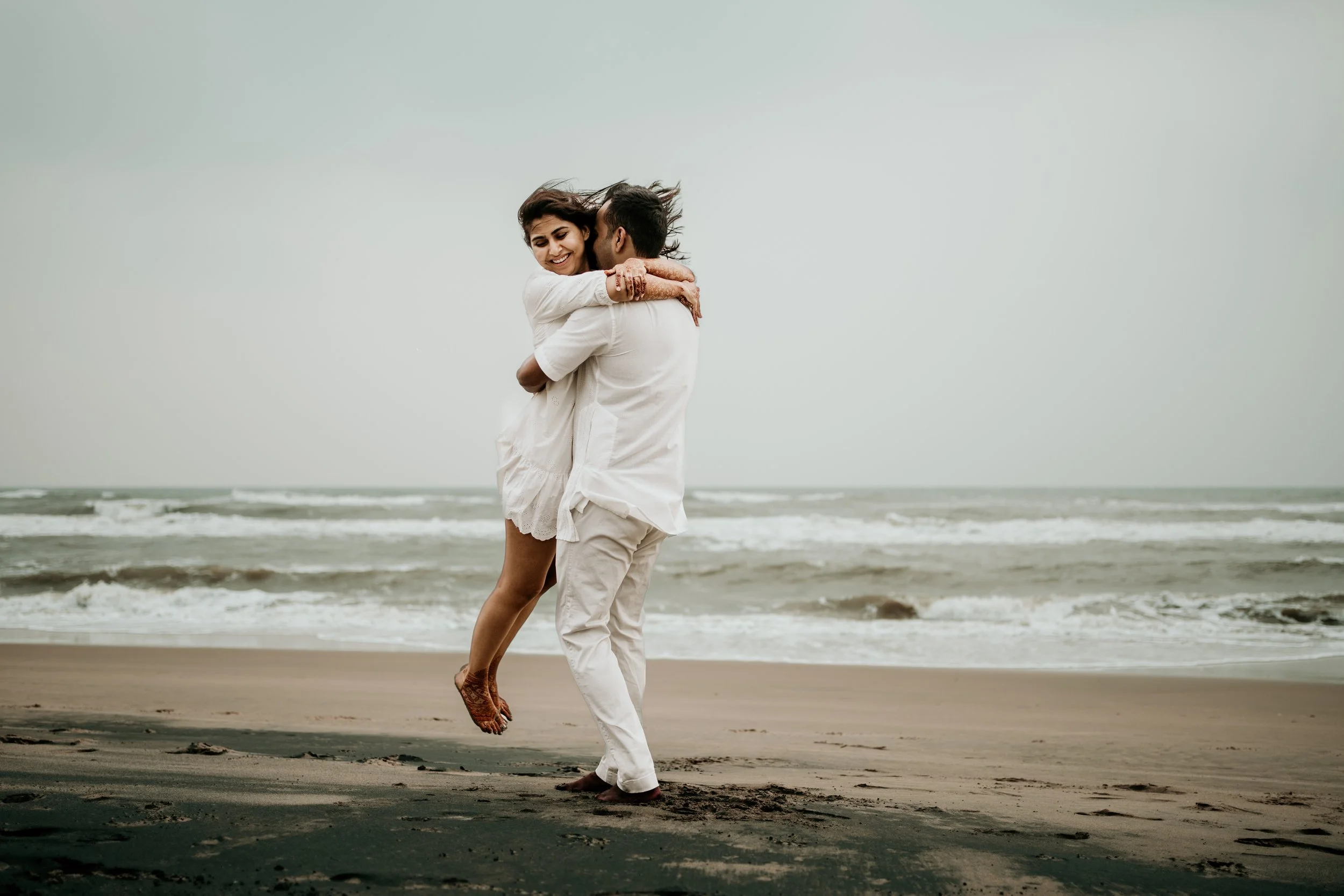 A couple embracing and dancing on a beach with waves in the background, both wearing white clothing.