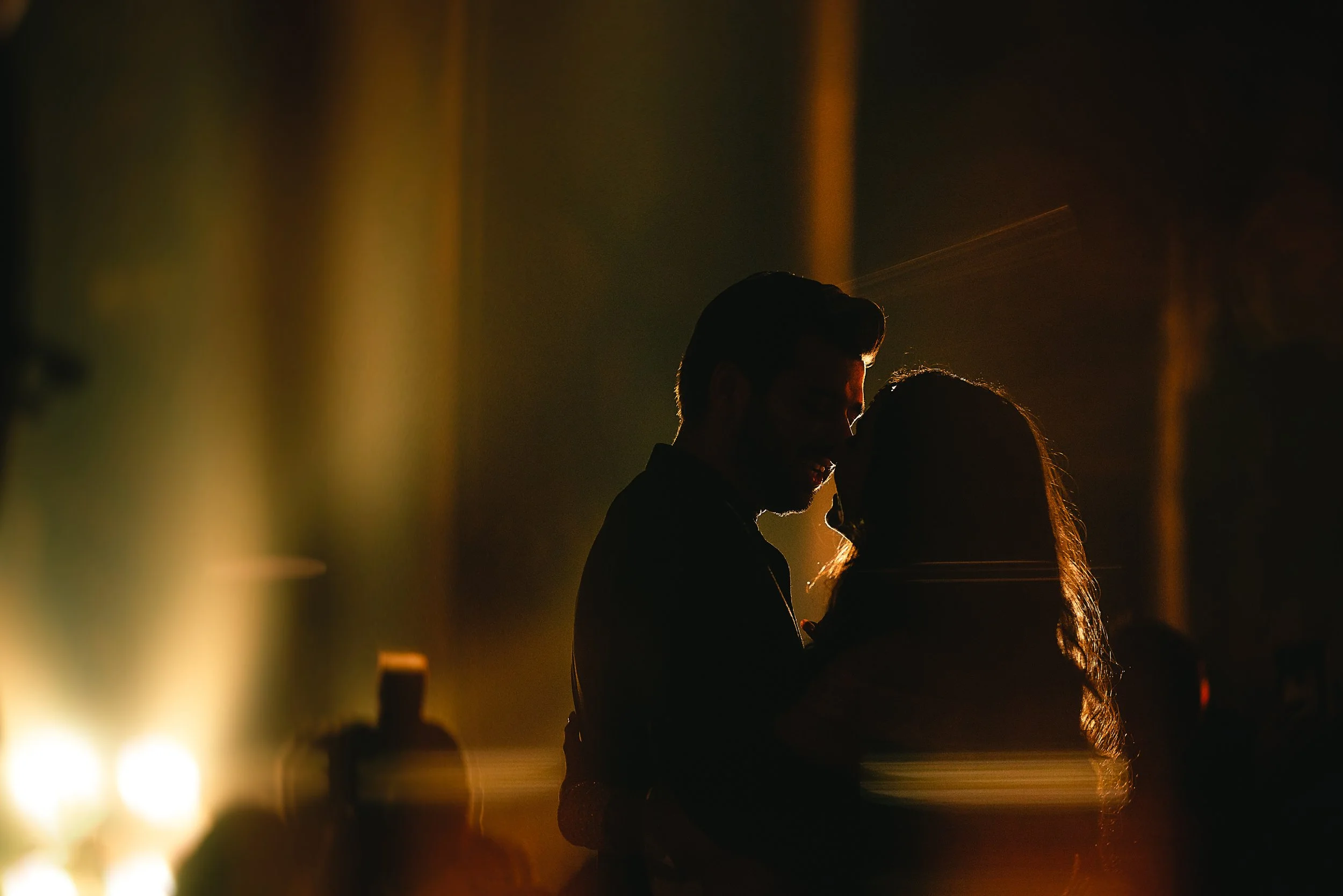 Silhouettes of a man and woman embracing and about to kiss at night, with warm lighting in the background.