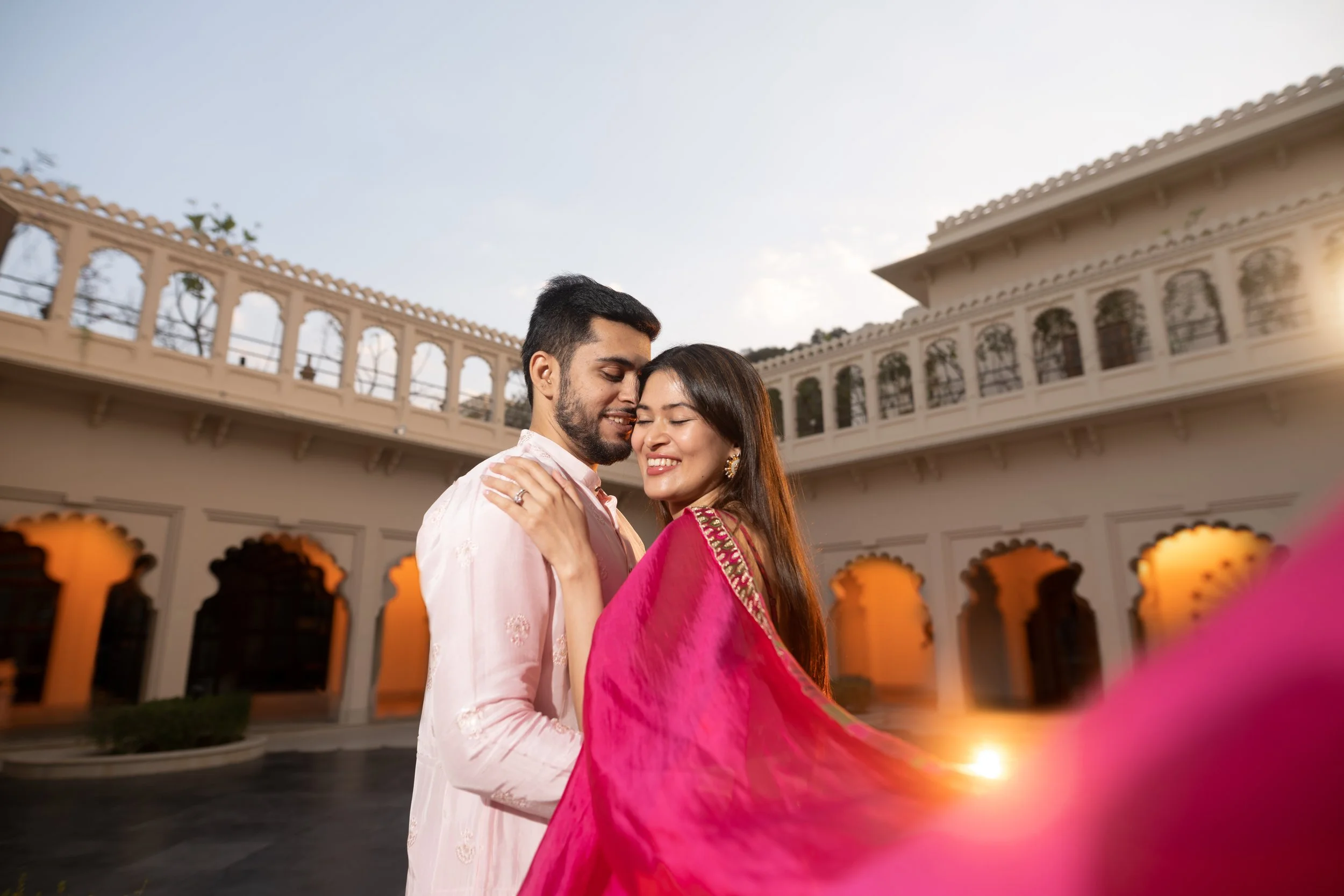 A happy couple dressed in traditional Indian attire embracing outdoors during sunset at a historic white building with arched windows and balconies.
